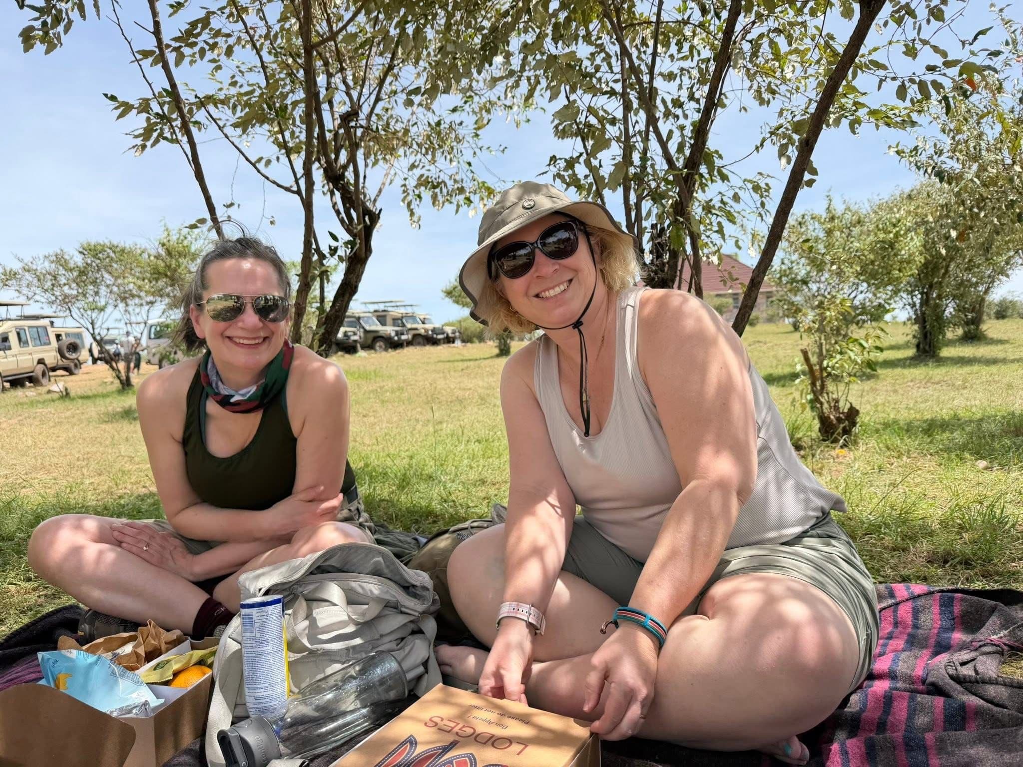 Picnic on the Maasai Mara with two travelers sitting on a blanket under a tree and safari vehicles in the background, Kenya.