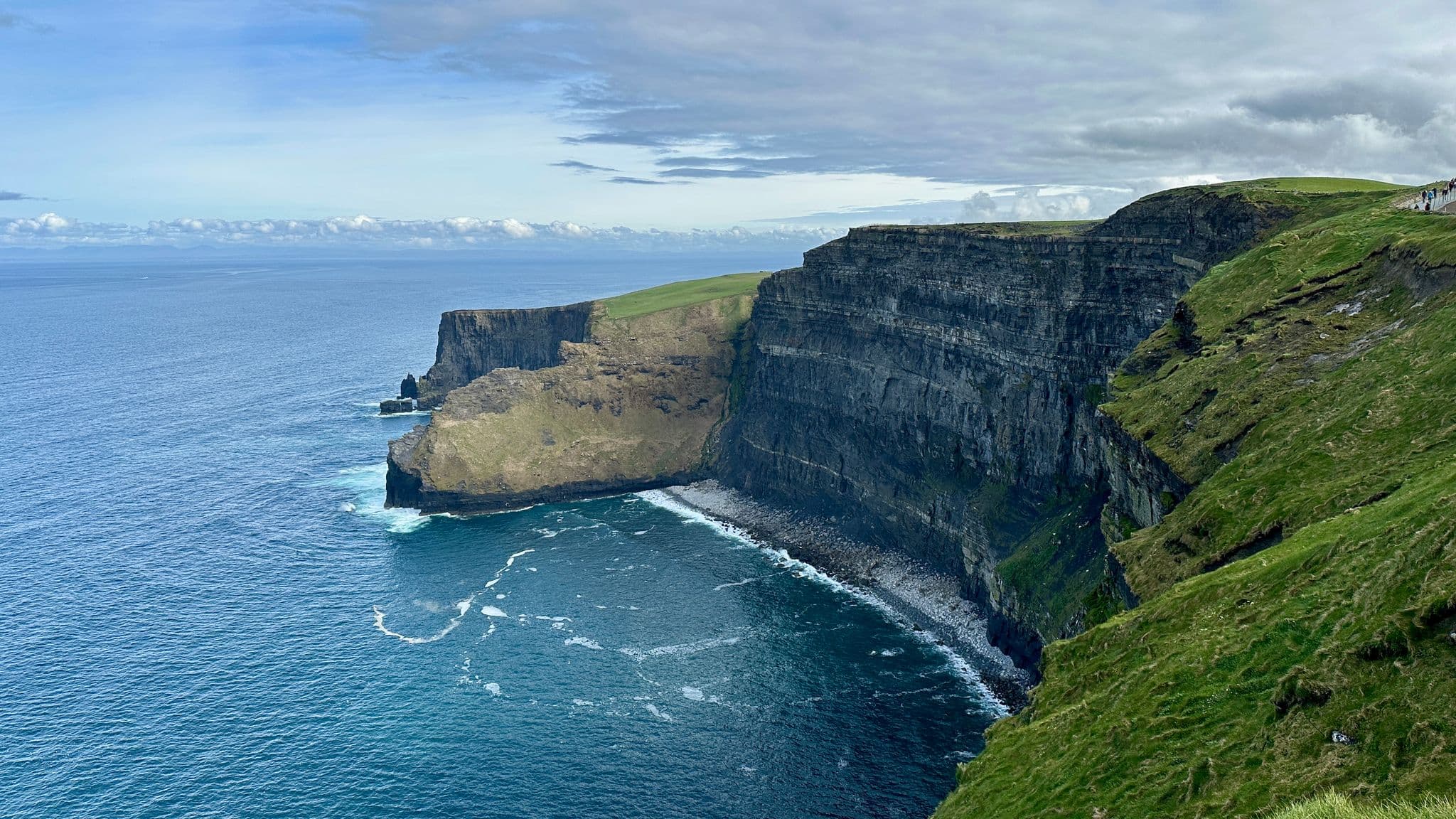 Cliffs of Moher towering above the Atlantic with waves below and visitors walking the grassy cliff path, County Clare, Ireland.