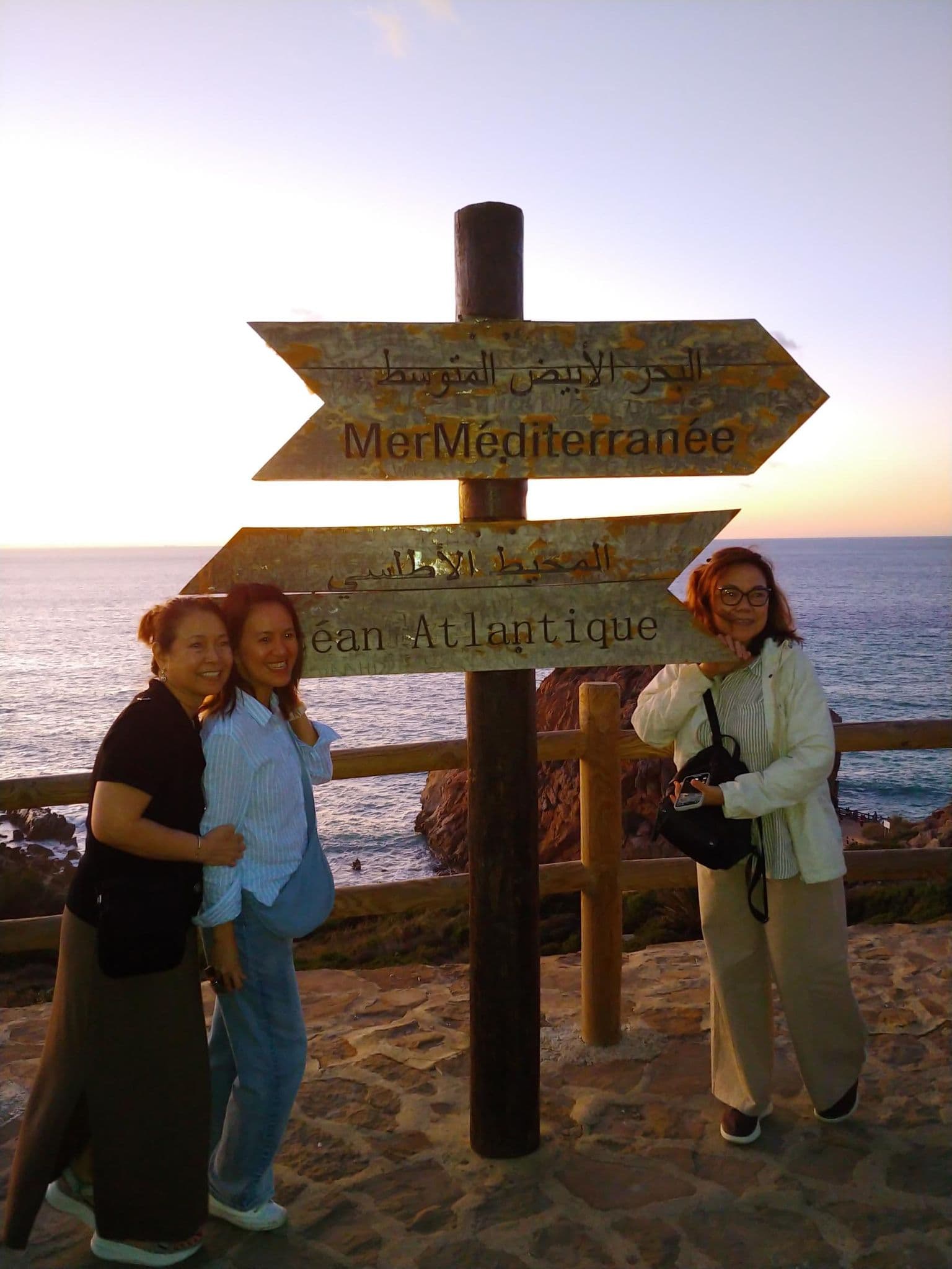 Wooden signpost reading "Mer Méditerranée" and "Océan Atlantique" at Cap Spartel, Tangier, Morocco, with three travelers by the sea.