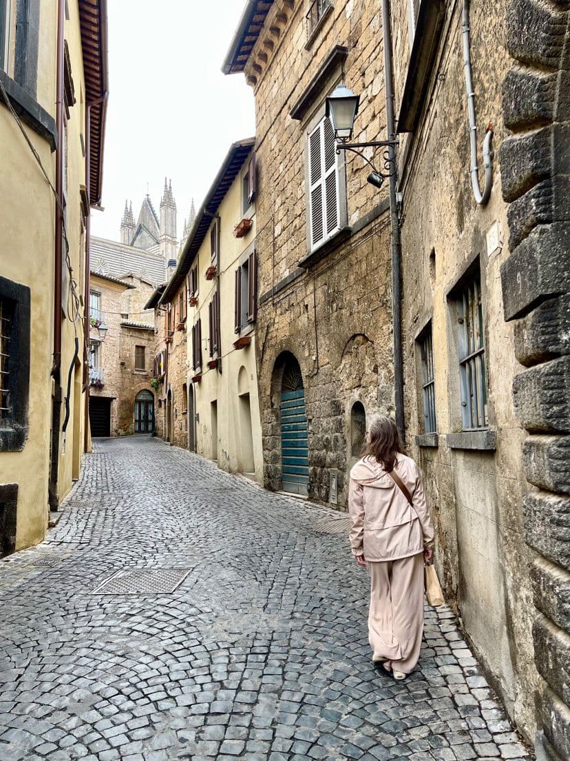 Duomo di Orvieto spires visible above a narrow cobblestone street as a woman walks away in Orvieto, Umbria, Italy.