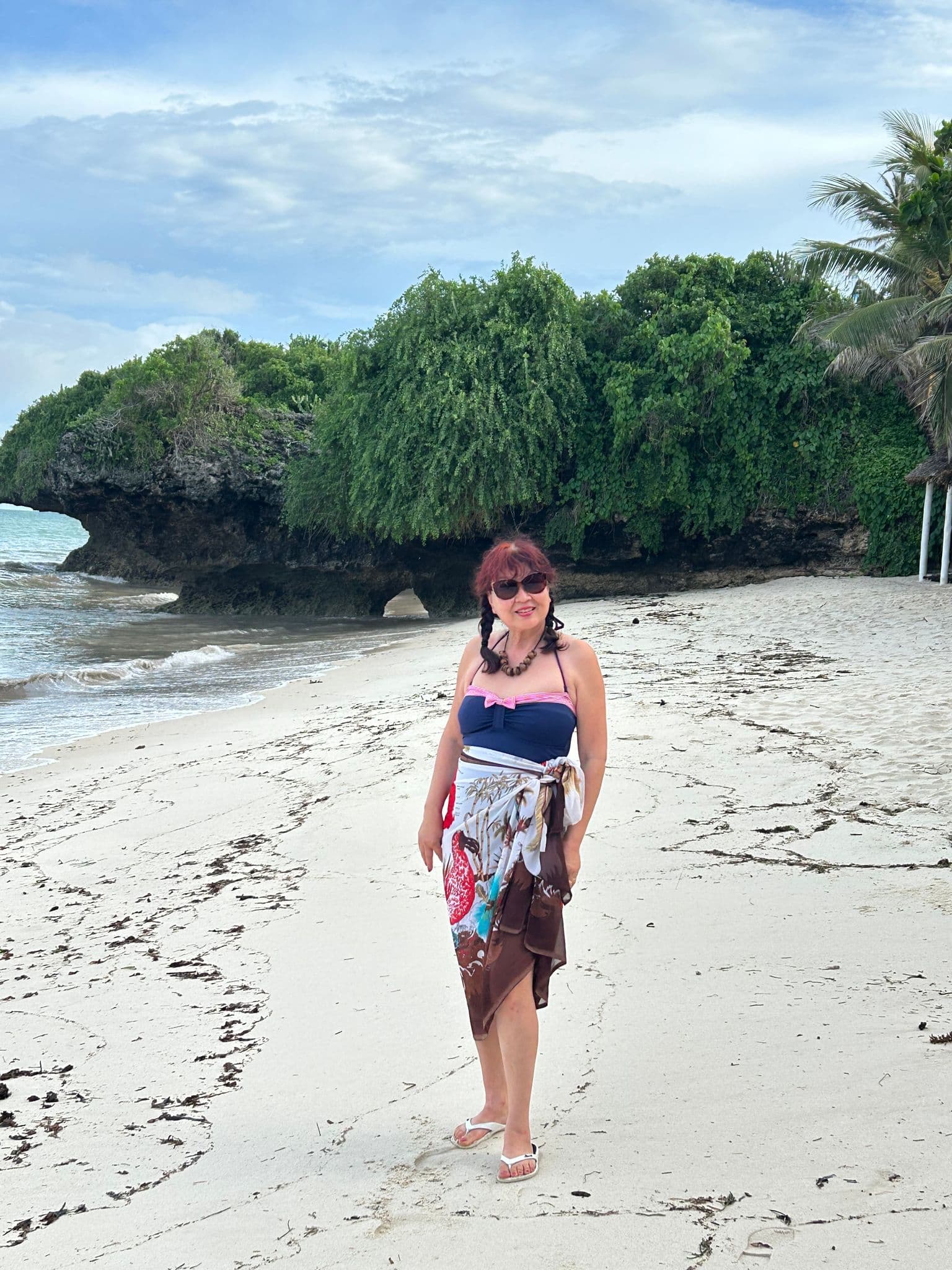 Diani Beach shoreline with a woman posing on the sand in front of a rocky, vegetation-covered sea cliff and palm trees, Kenya.