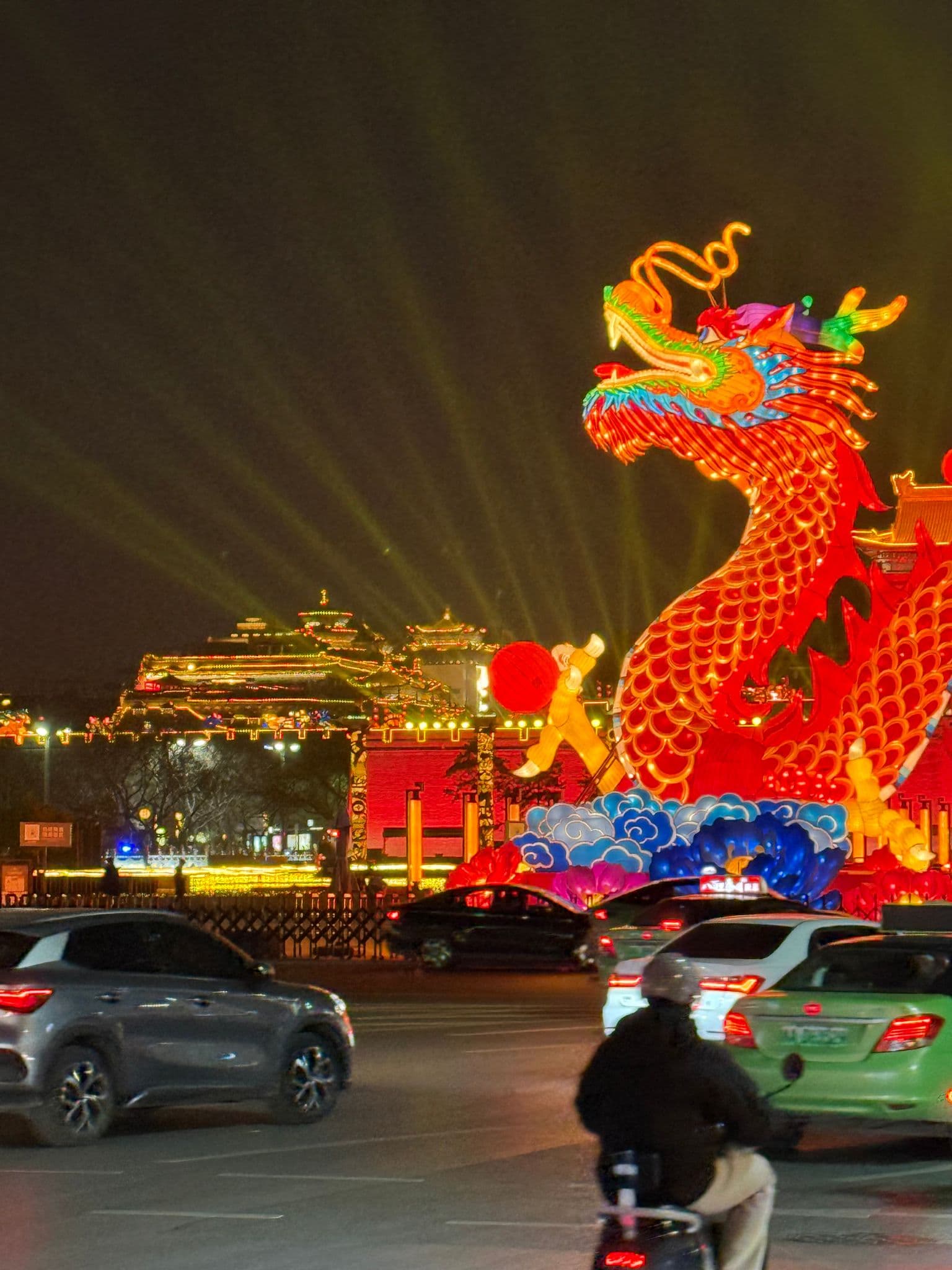 Giant illuminated dragon lantern at Bell Tower plaza in Xi'an, China, lit at night with traffic and a scooter passing in the foreground.
