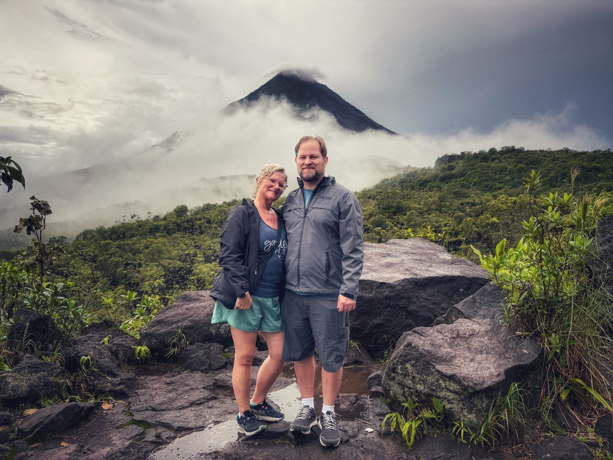 Arenal Volcano shrouded in mist with a couple posing on a rocky viewpoint, La Fortuna, Costa Rica.