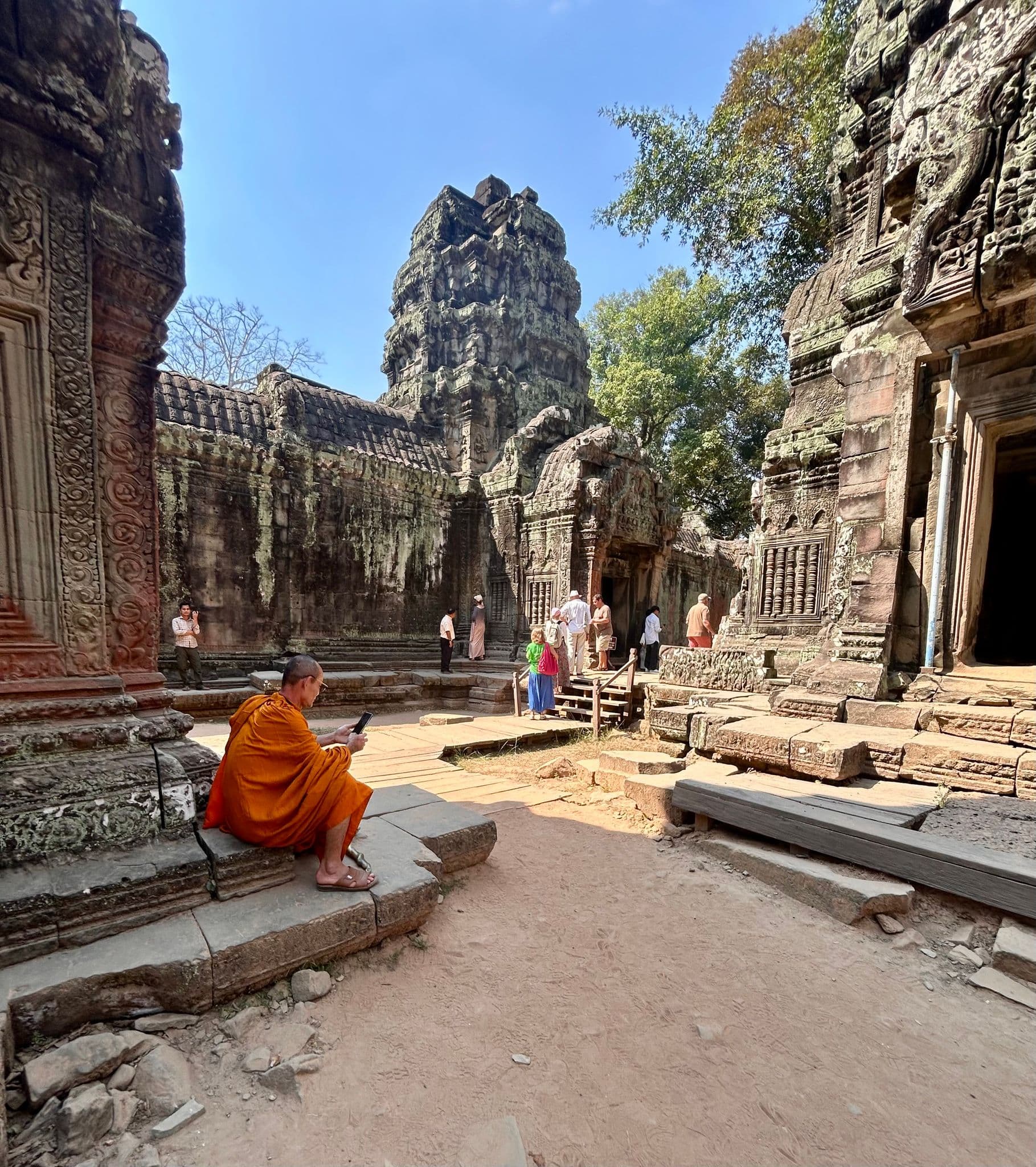 Ta Prohm temple ruins with a Buddhist monk in orange robes seated checking a phone, visitors nearby, Angkor, Cambodia.