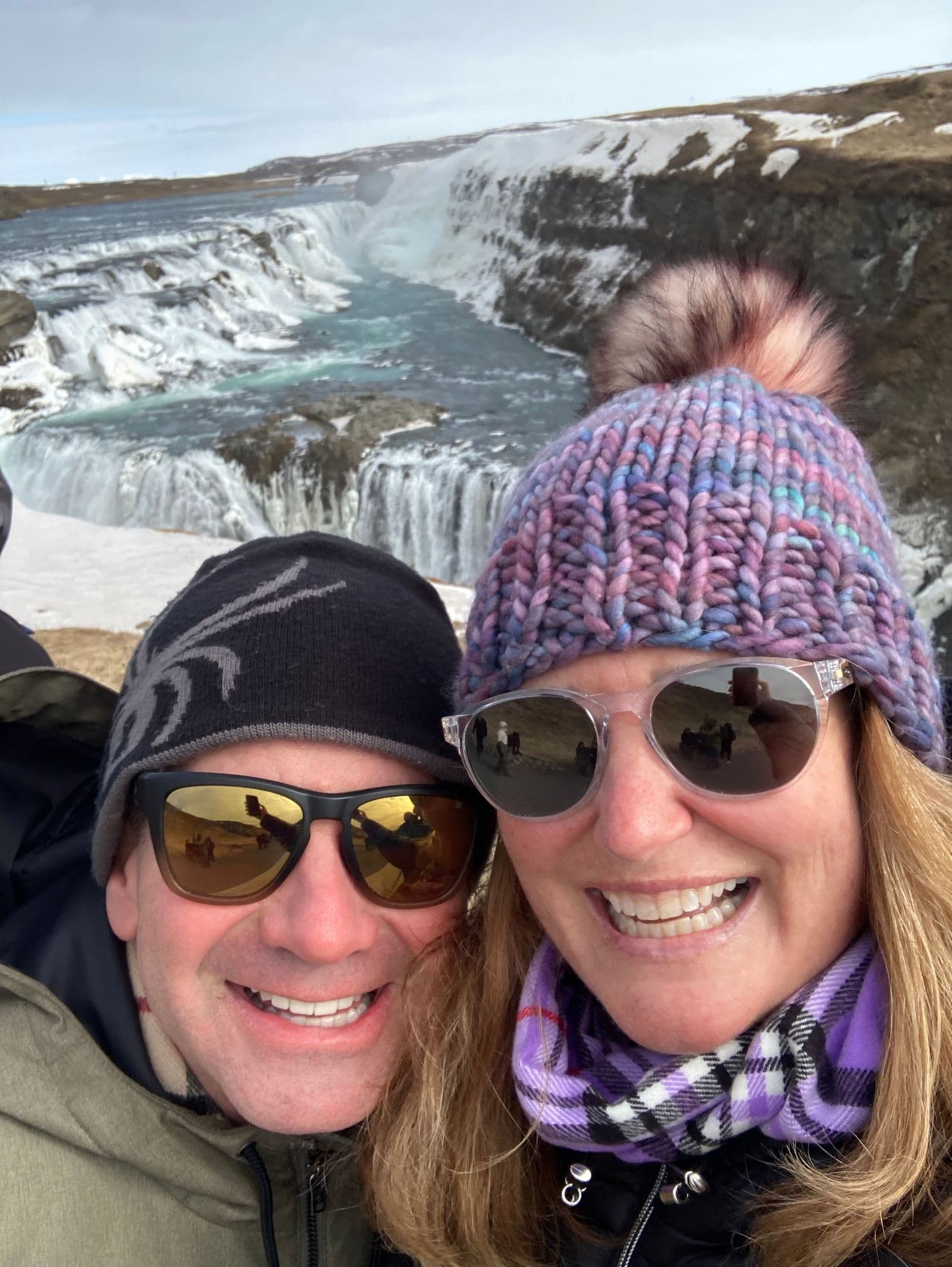 Gullfoss waterfall in Iceland with two travelers taking a close-up selfie wearing winter hats and sunglasses.