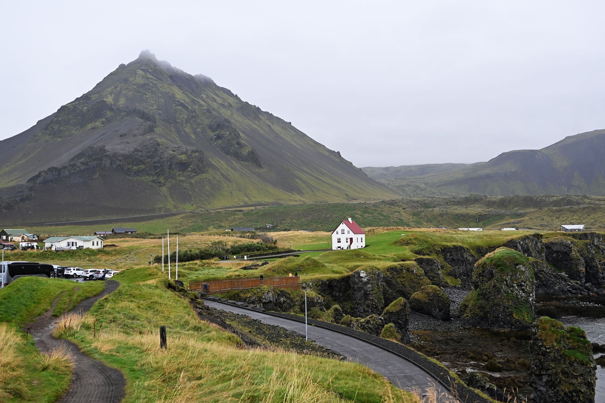 Cliffs at Arnarstapi with a small white house and a winding coastal path beneath a steep mountain in Iceland.