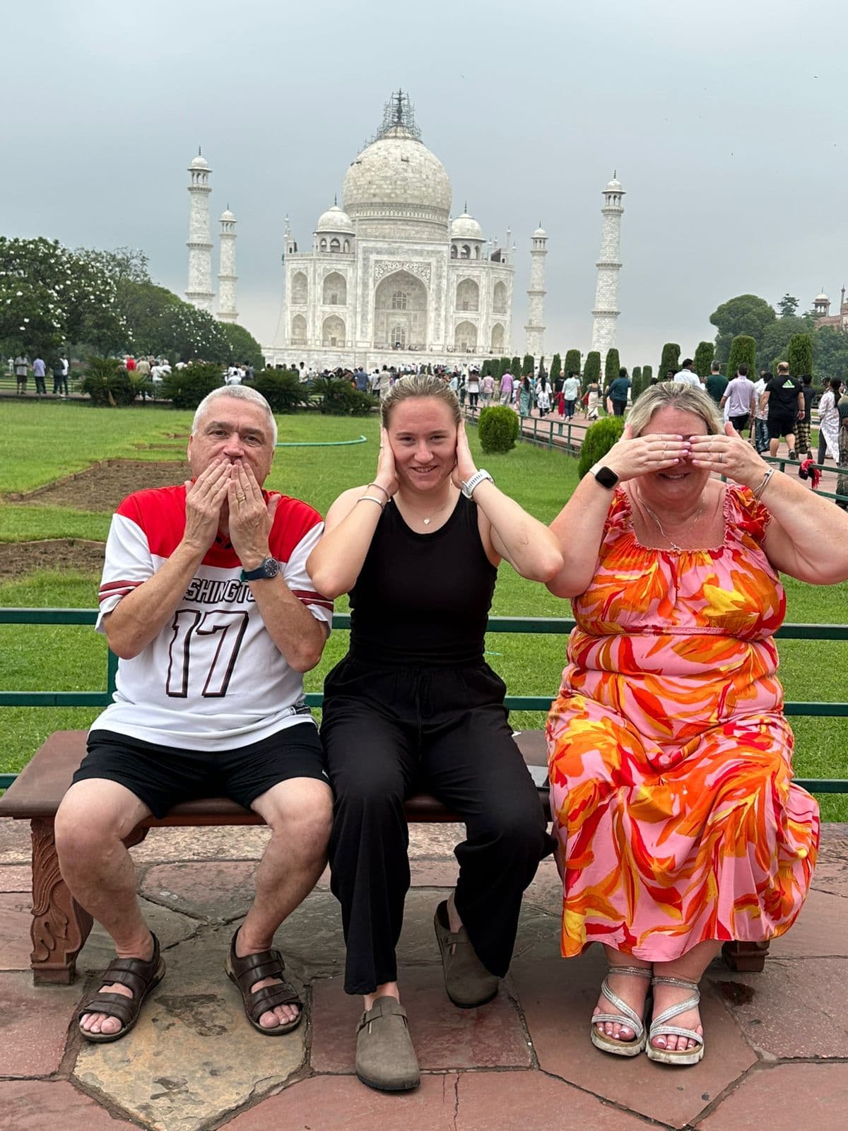 Taj Mahal with three travelers seated on a bench in front, each covering mouth, ears, or eyes, Agra, India.