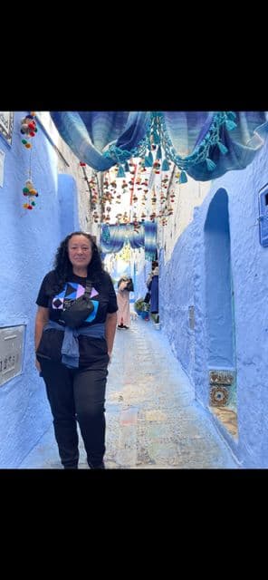 Blue alley of Chefchaouen with a woman standing in the foreground beneath hanging textiles and ornaments, Chefchaouen, Morocco.