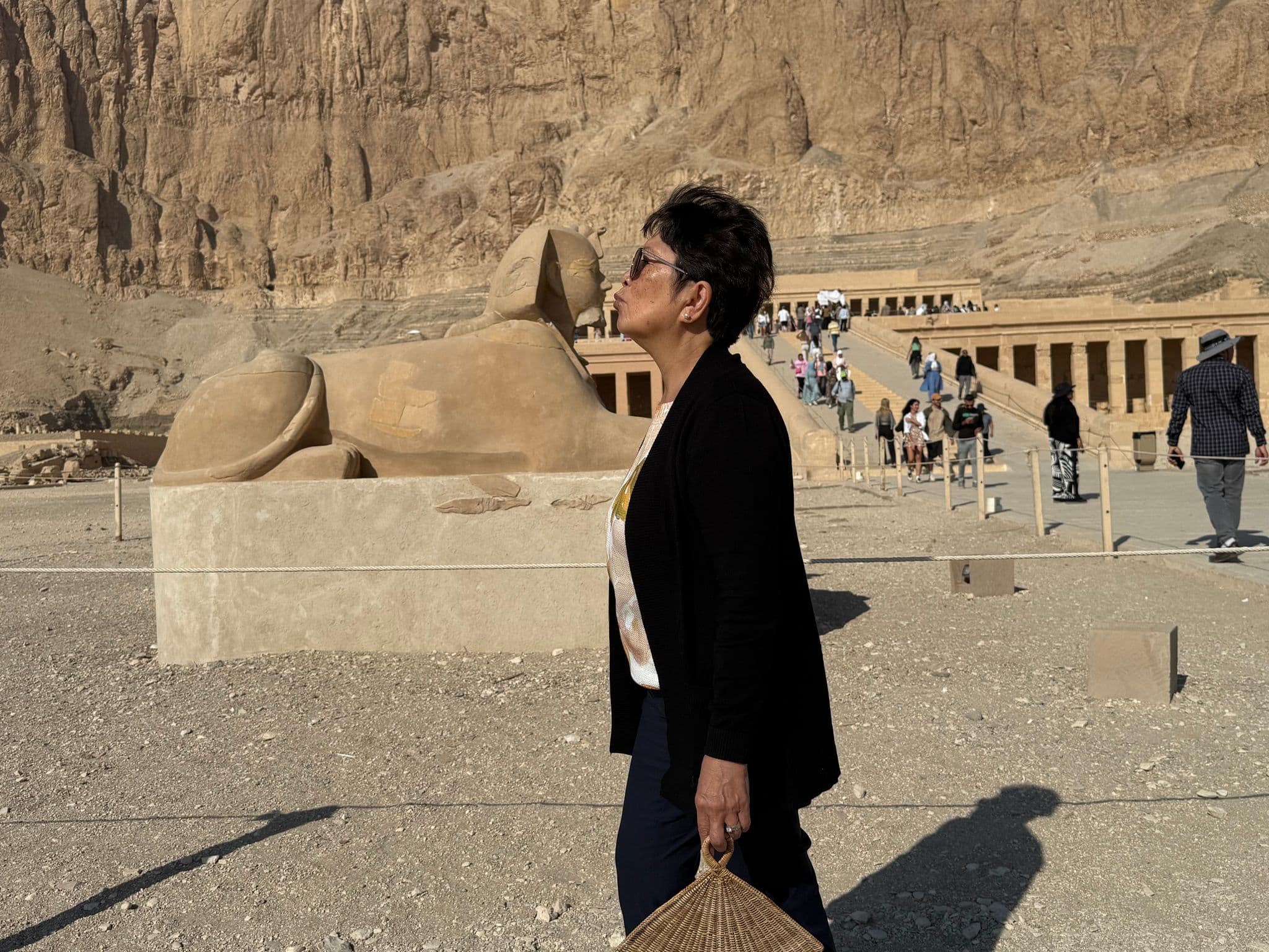 Mortuary Temple of Hatshepsut at Deir el-Bahari, Egypt, with a visitor standing in profile beside a sandstone sphinx.