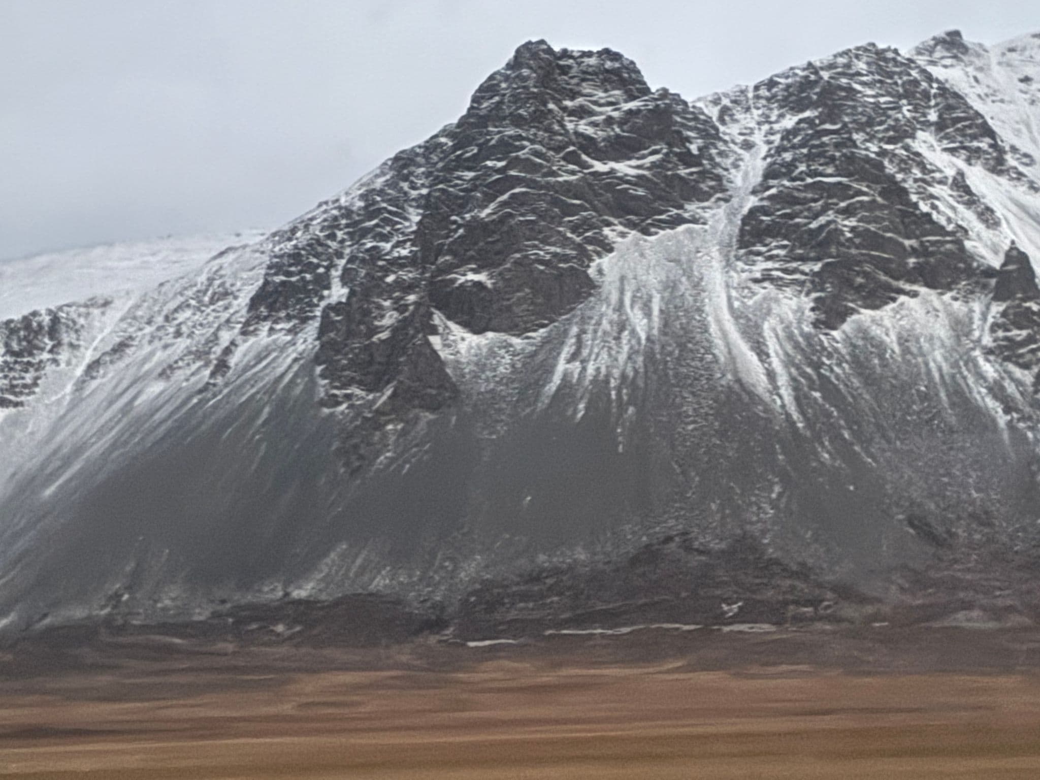 Snow-dusted rocky mountain rising above a flat brown plain under a pale morning sky.