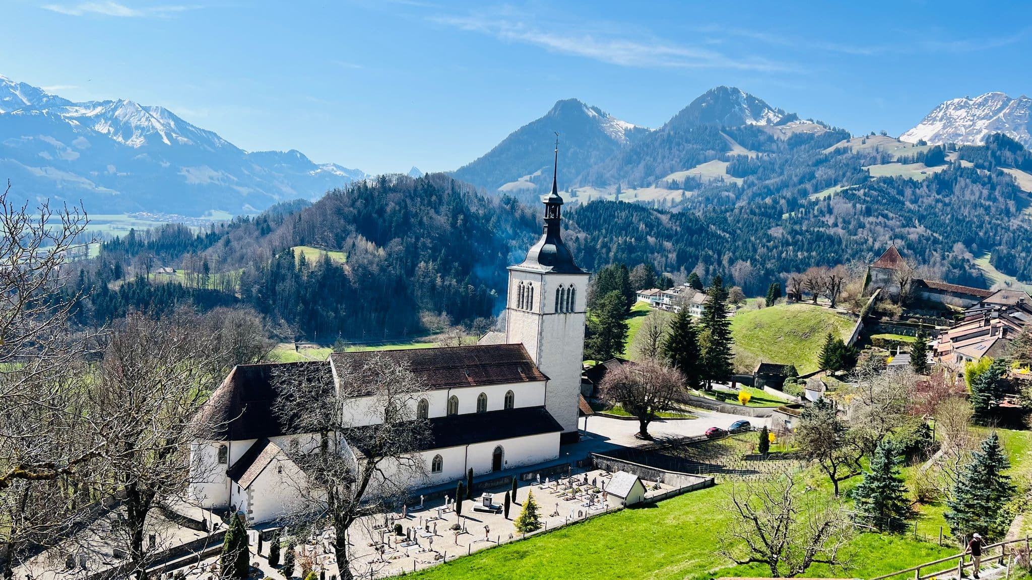 Gruyères Castle and a white church with a cemetery on a green hillside, alpine peaks behind, Gruyères, Switzerland.