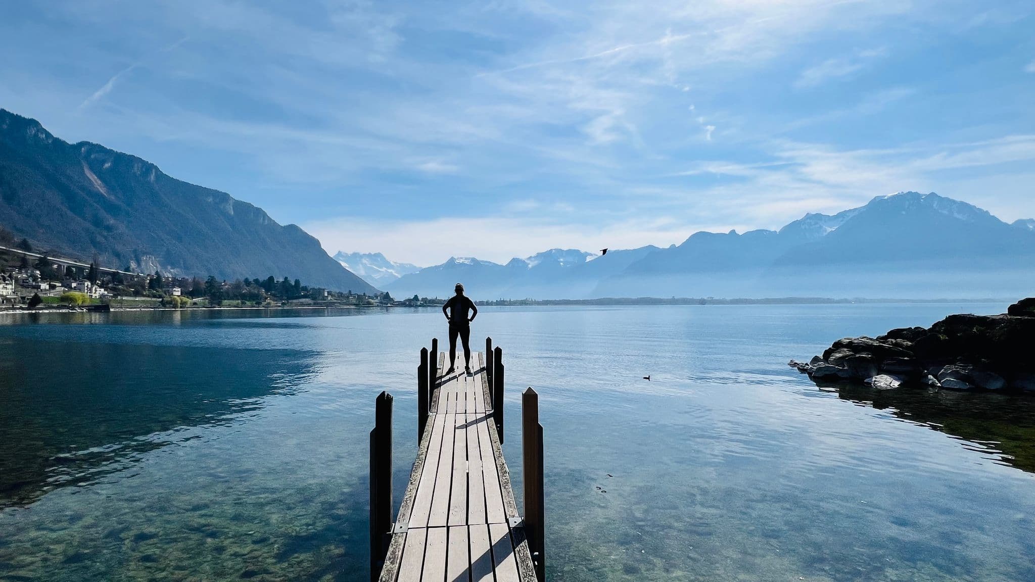 Person standing at the end of a wooden pier looking across a clear alpine lake toward the Swiss Alps in Switzerland.