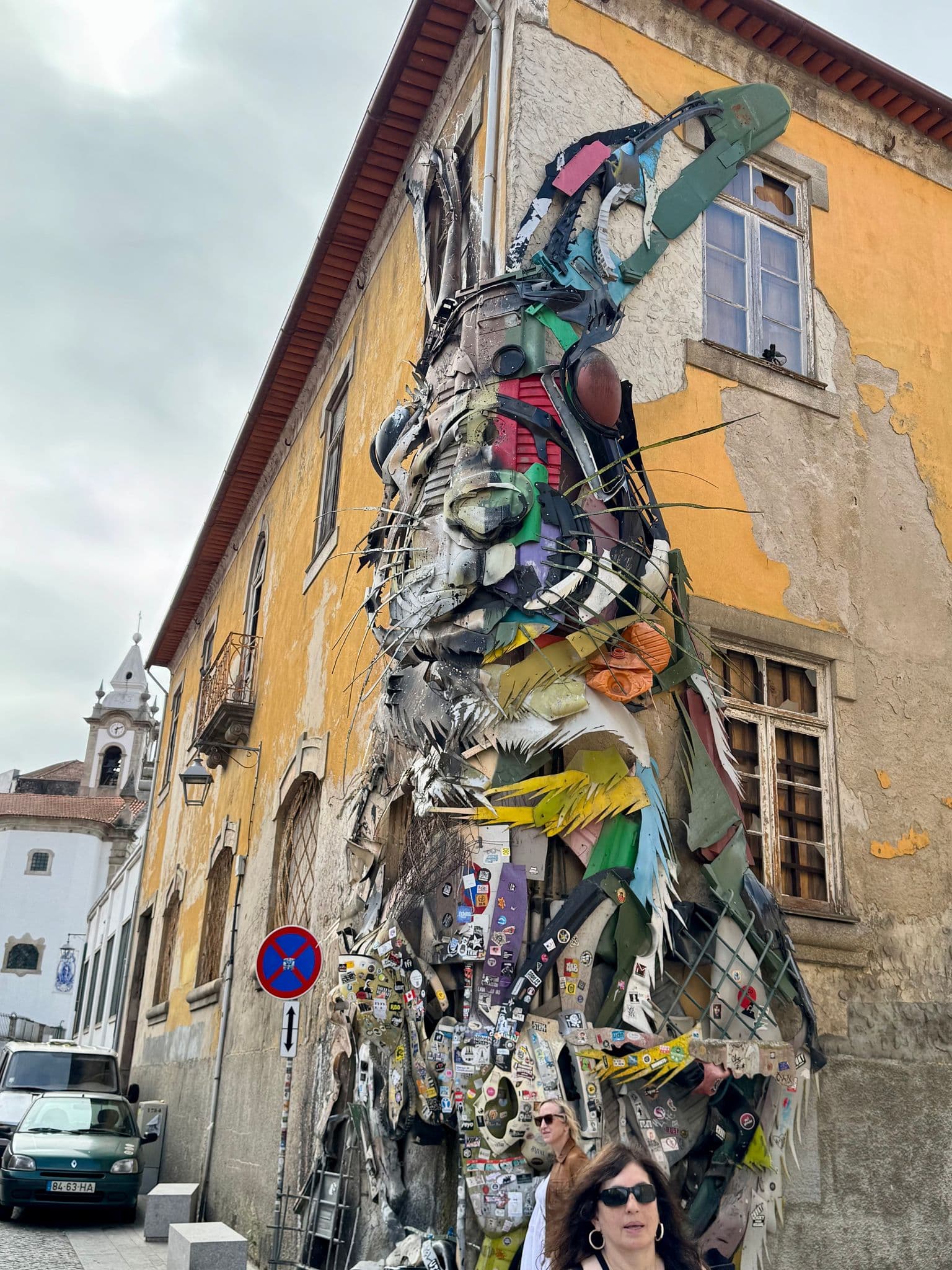 Large recycled-material rabbit sculpture mounted on a yellow building, with pedestrians on a narrow street in Portugal.