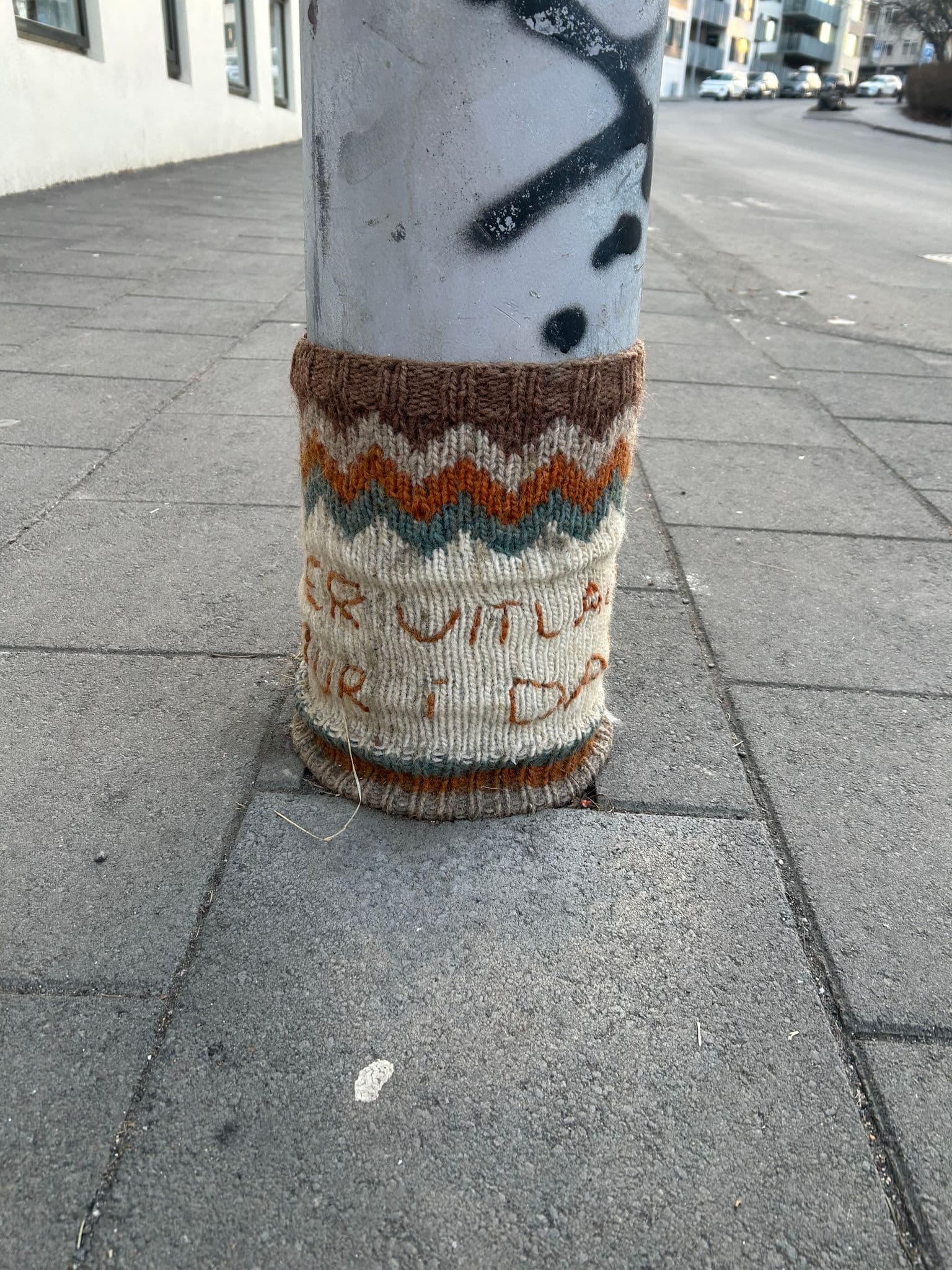 Lamp post wrapped in a knitted cozy on a Reykjavík sidewalk, viewed from ground level with paving stones, Iceland.