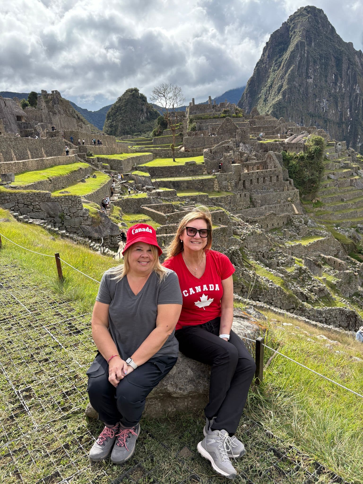Machu Picchu ruins with two women sitting on a rock in the foreground wearing Canada shirts and a Canada hat, Cusco, Peru.