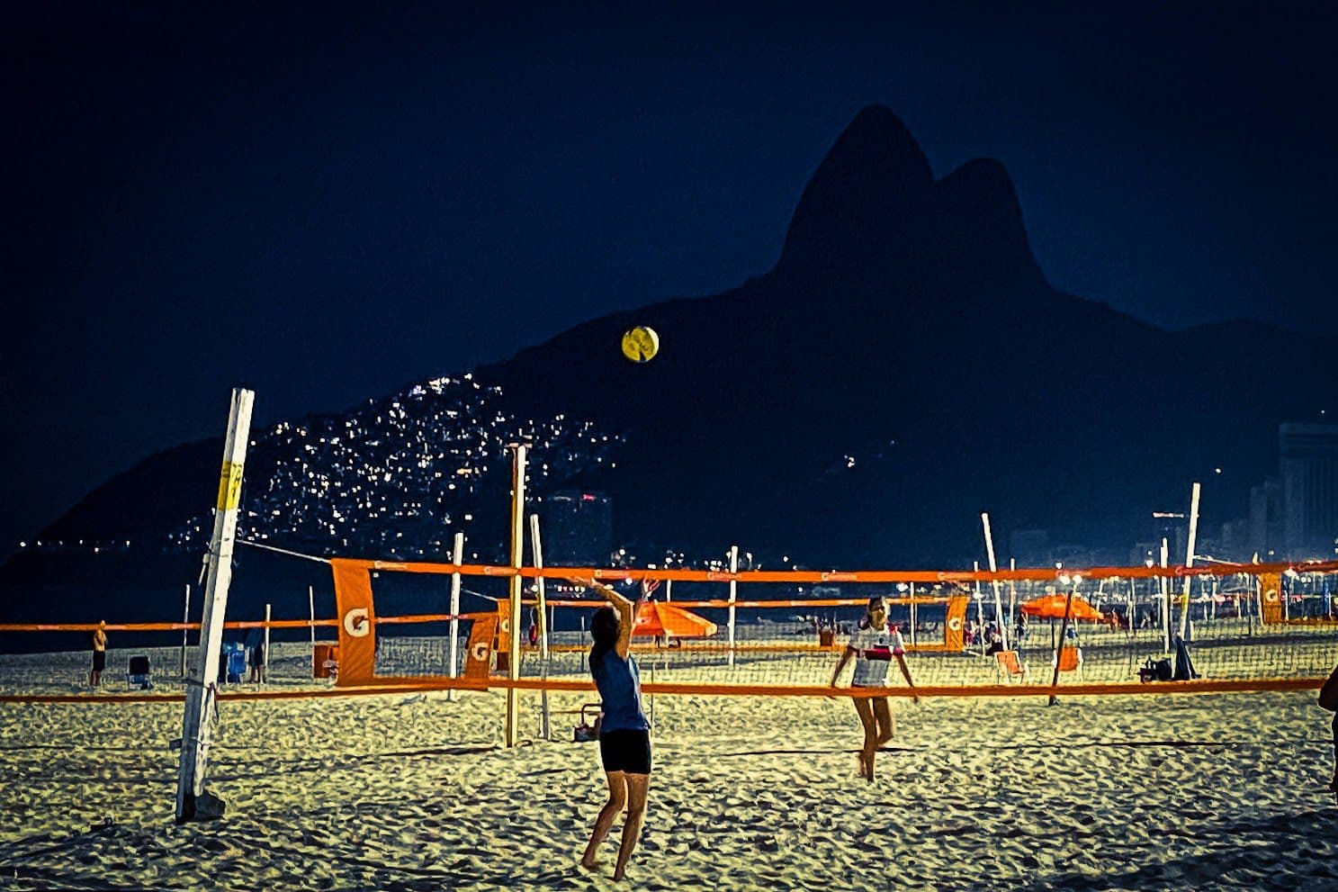 Ipanema Beach volleyball nets at night with Morro Dois Irmãos silhouetted and players jumping on the sand, Rio de Janeiro, Brazil.