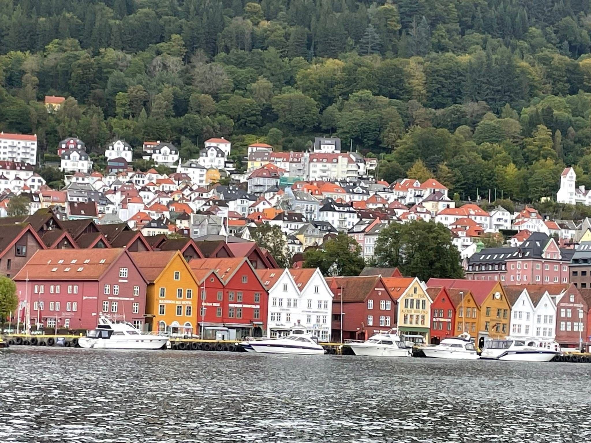 Colorful wooden houses of Bryggen waterfront lined along the harbor with boats moored and a forested hillside behind, Bergen, Norway.