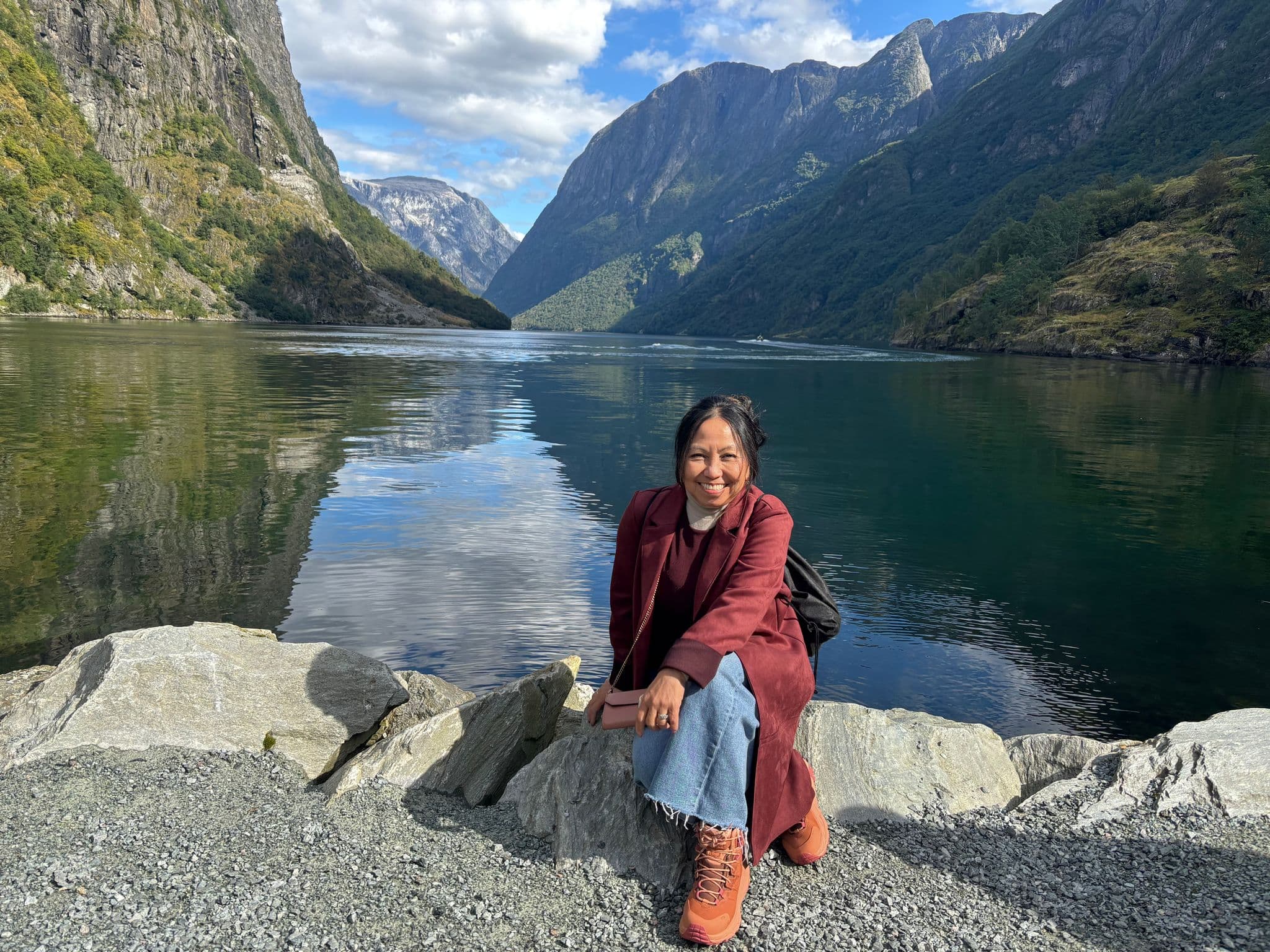 Woman sitting on rocks by a fjord with steep mountains reflected in the water, Norway.
