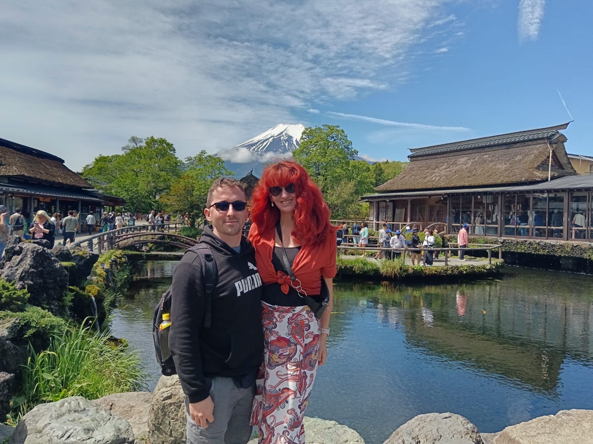 Mount Fuji rising behind a traditional pond at Oshino Hakkai, Japan, with a couple posing at the water's edge.