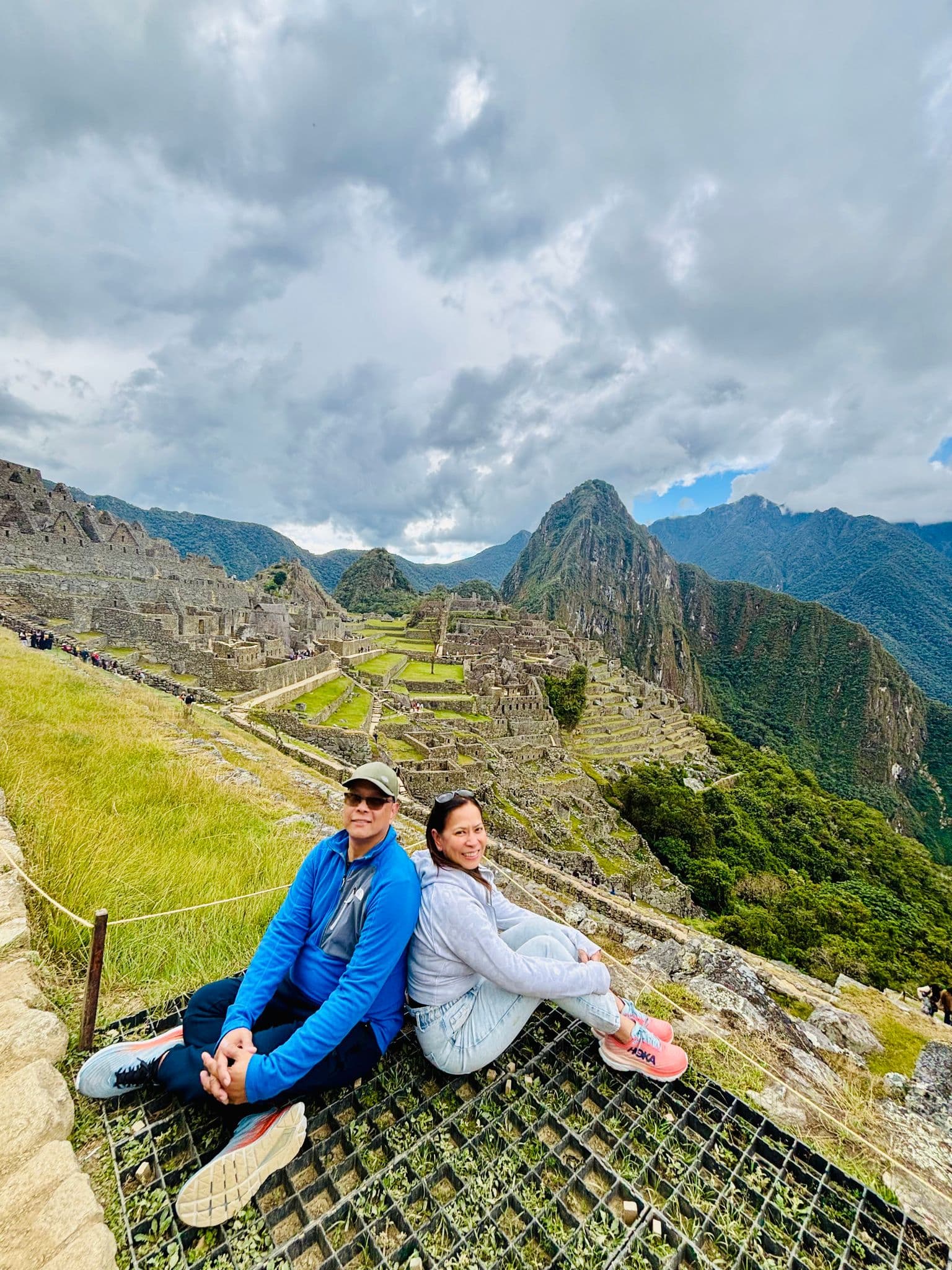 Machu Picchu ruins with two travelers sitting on a grassy terrace, Huayna Picchu and mountain terraces visible, Cusco Region, Peru
