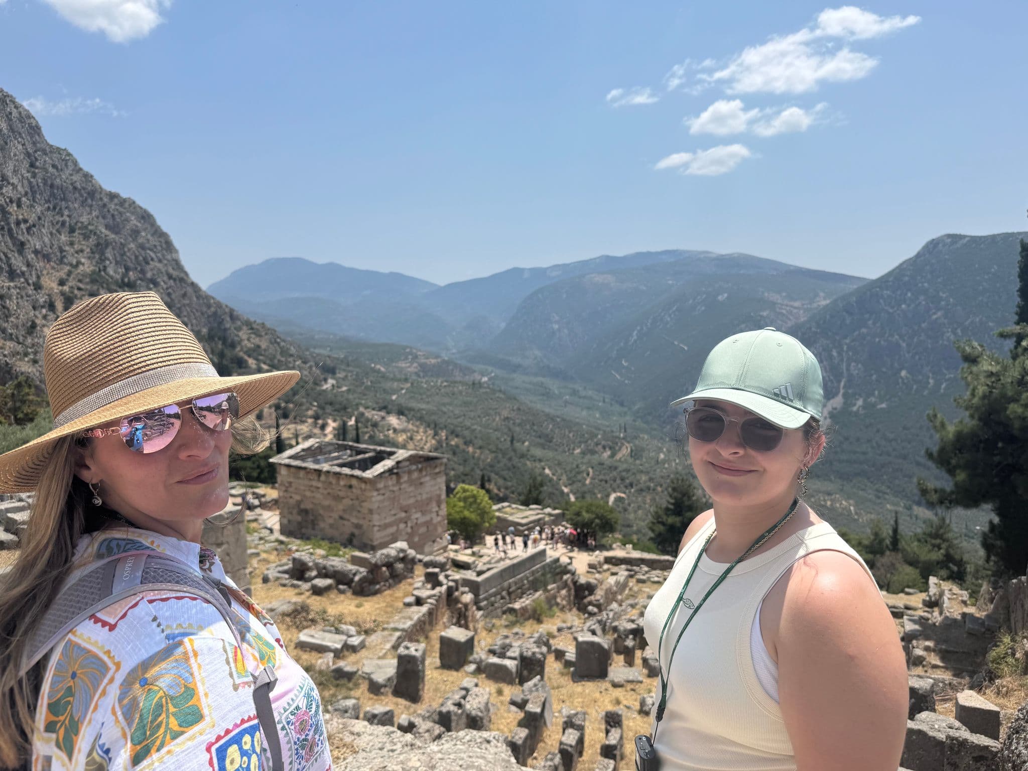 Temple ruins at Delphi with two travelers posing at an overlook and a mountainous valley behind, Delphi, Greece.