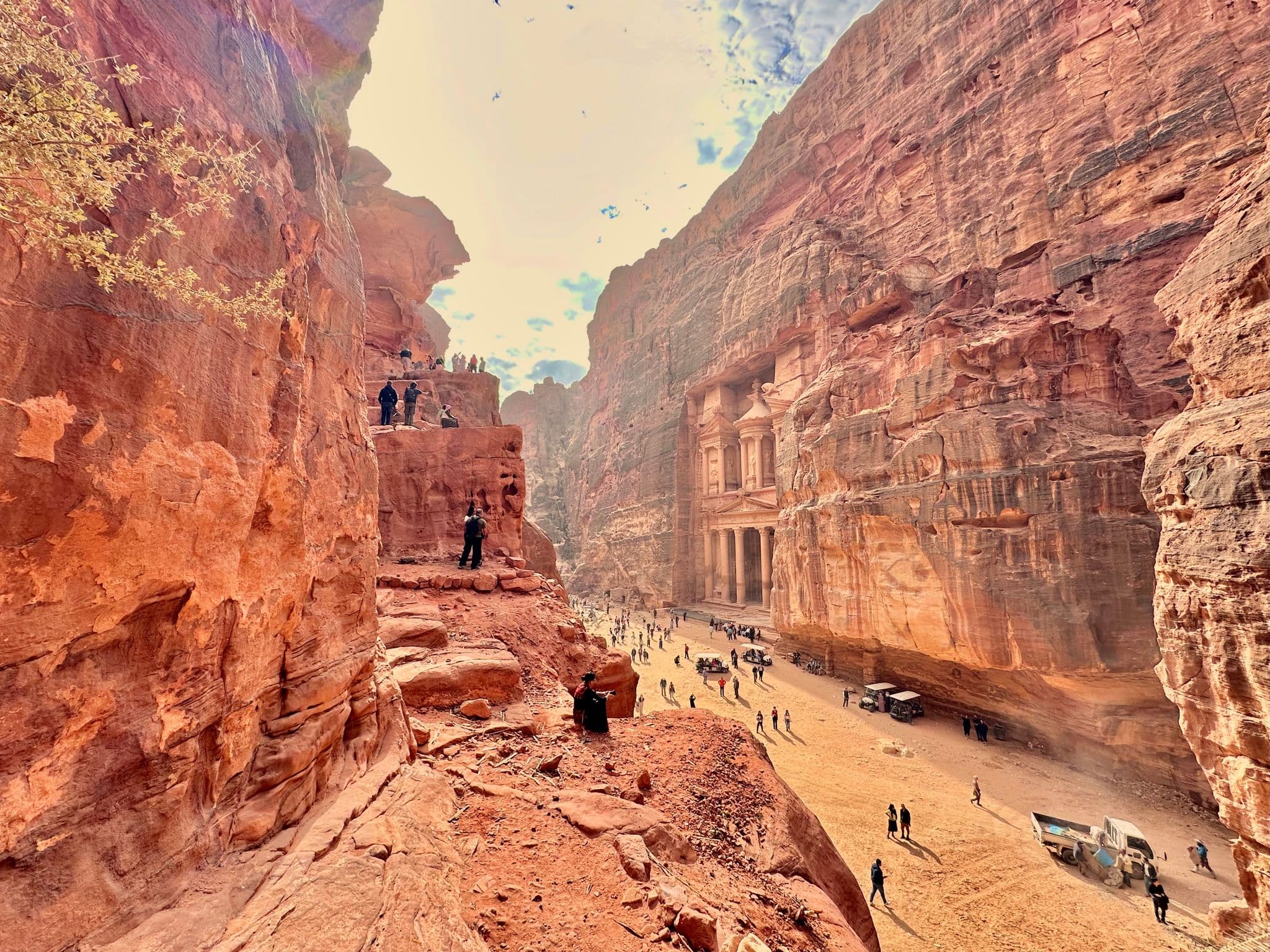 Al-Khazneh (The Treasury) in Petra framed by red sandstone cliffs with tourists climbing rocks and walking in the canyon, Jordan.