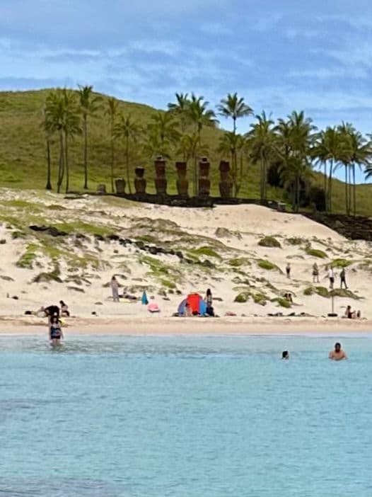 Moai statues at Anakena Beach standing on the sand with people swimming and relaxing in the foreground, Easter Island, Chile.