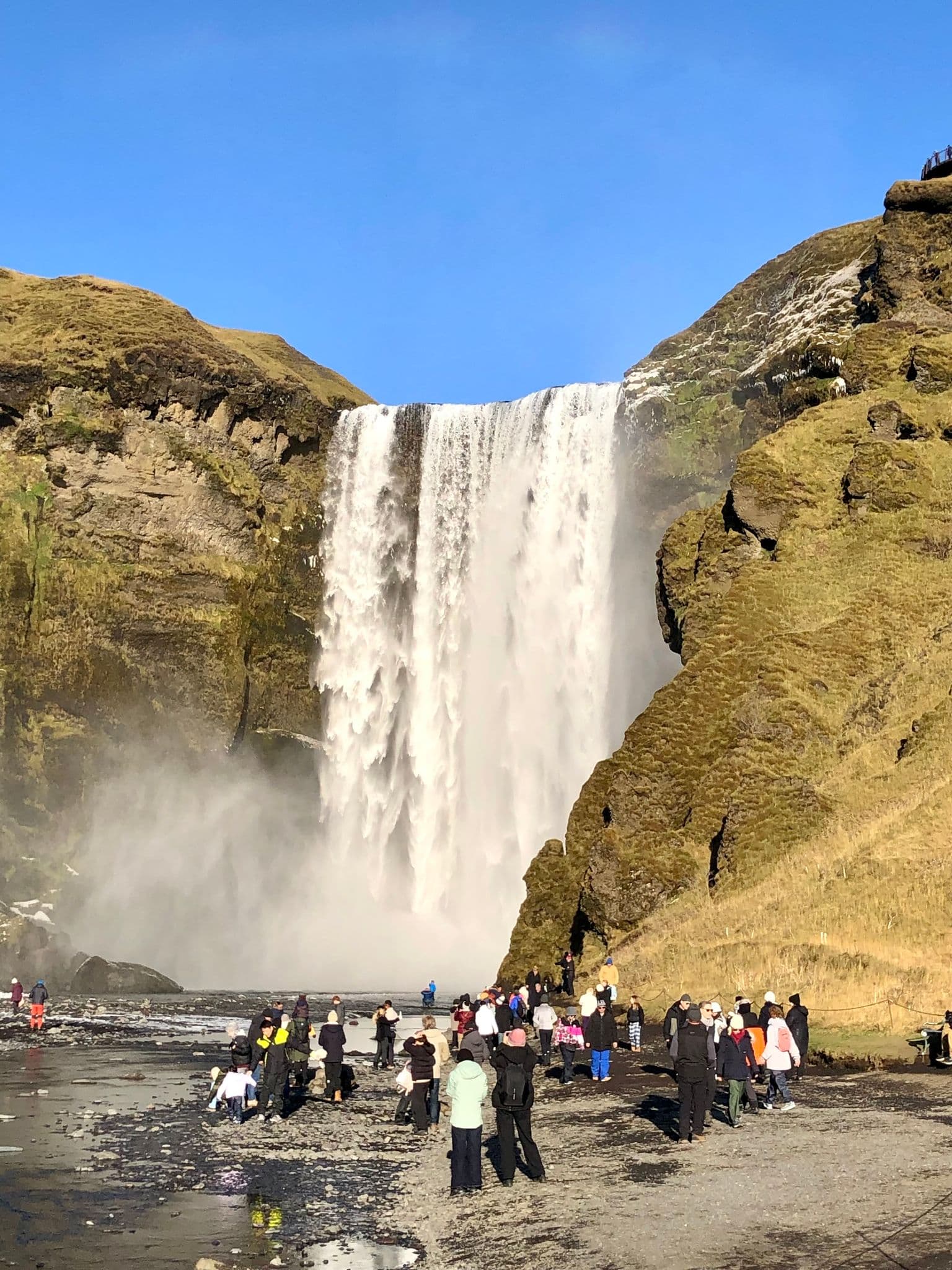 Skógafoss waterfall cascading over a mossy cliff with tourists standing and photographing near the base in Skógar, Iceland.