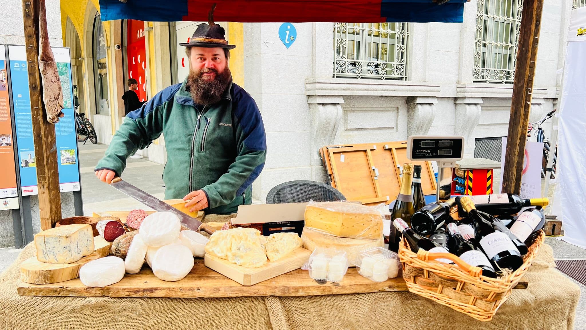 Market vendor slicing cheese behind a stall of assorted cheeses and wine bottles at an outdoor street market, Spain.