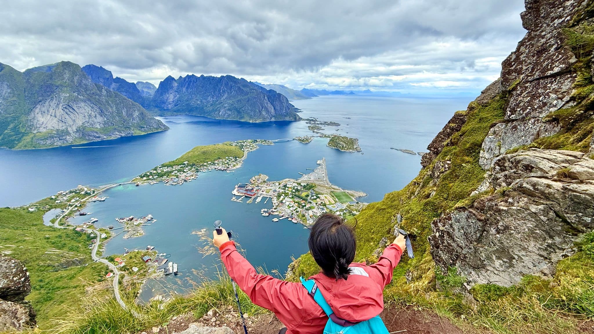Reinebringen viewpoint with a hiker holding trekking poles overlooking Reine village and surrounding fjords in Lofoten, Norway.