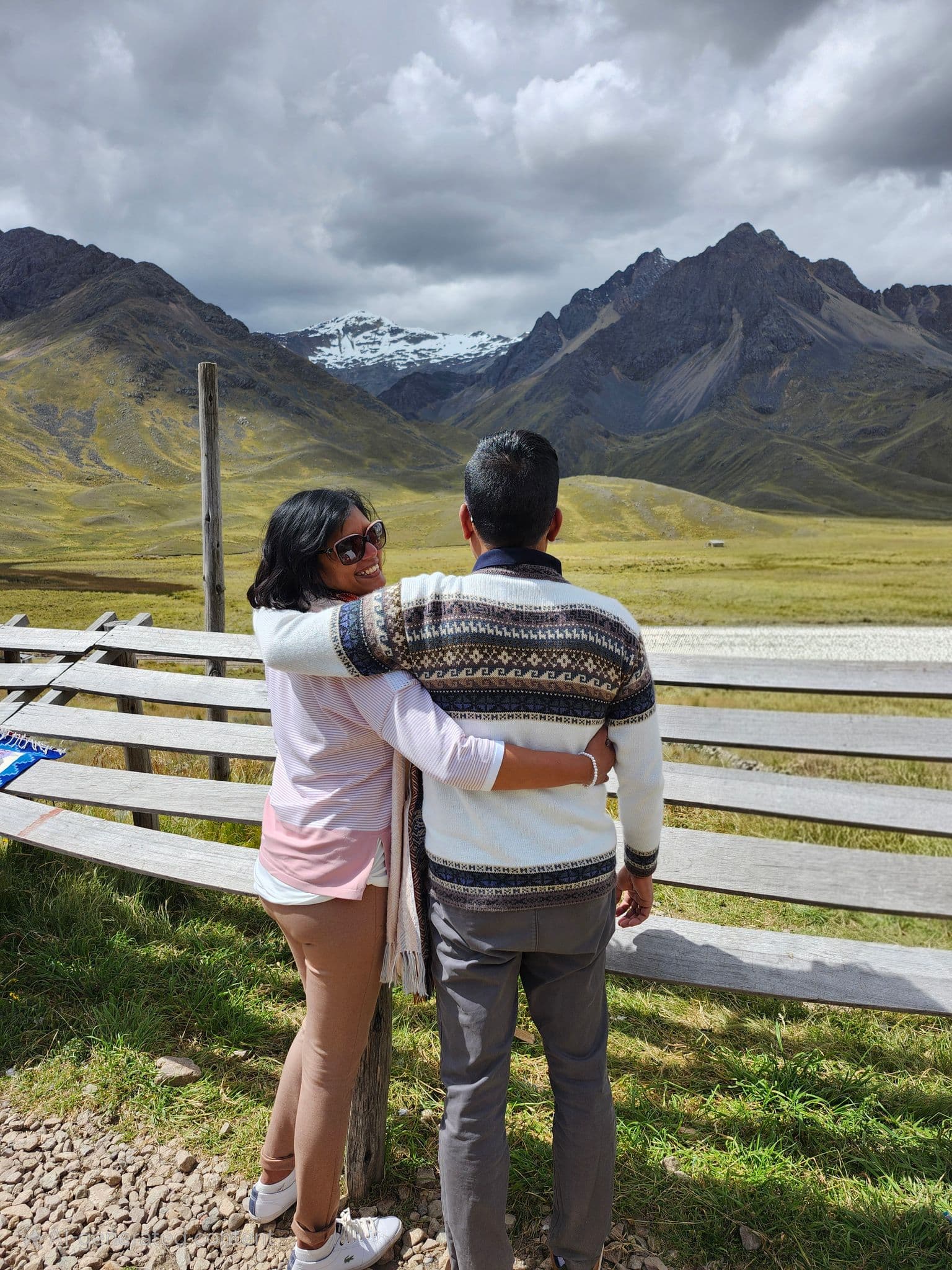 Couple embracing by a wooden fence at La Raya Pass with snow-capped Andes mountains in Peru.