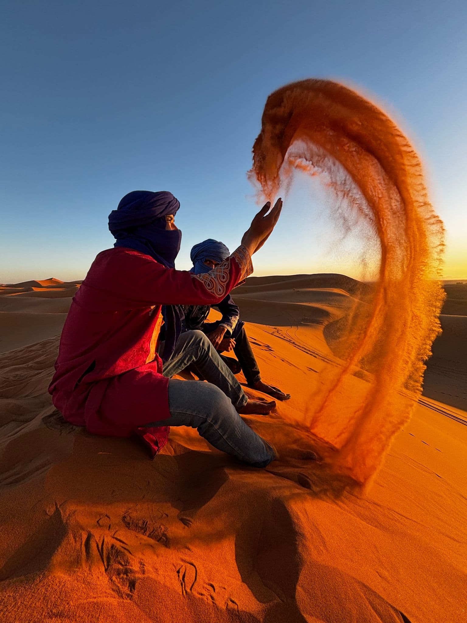 Two men in blue turbans sitting on a dune as one throws a large arc of sand on the Sahara Desert at sunset.