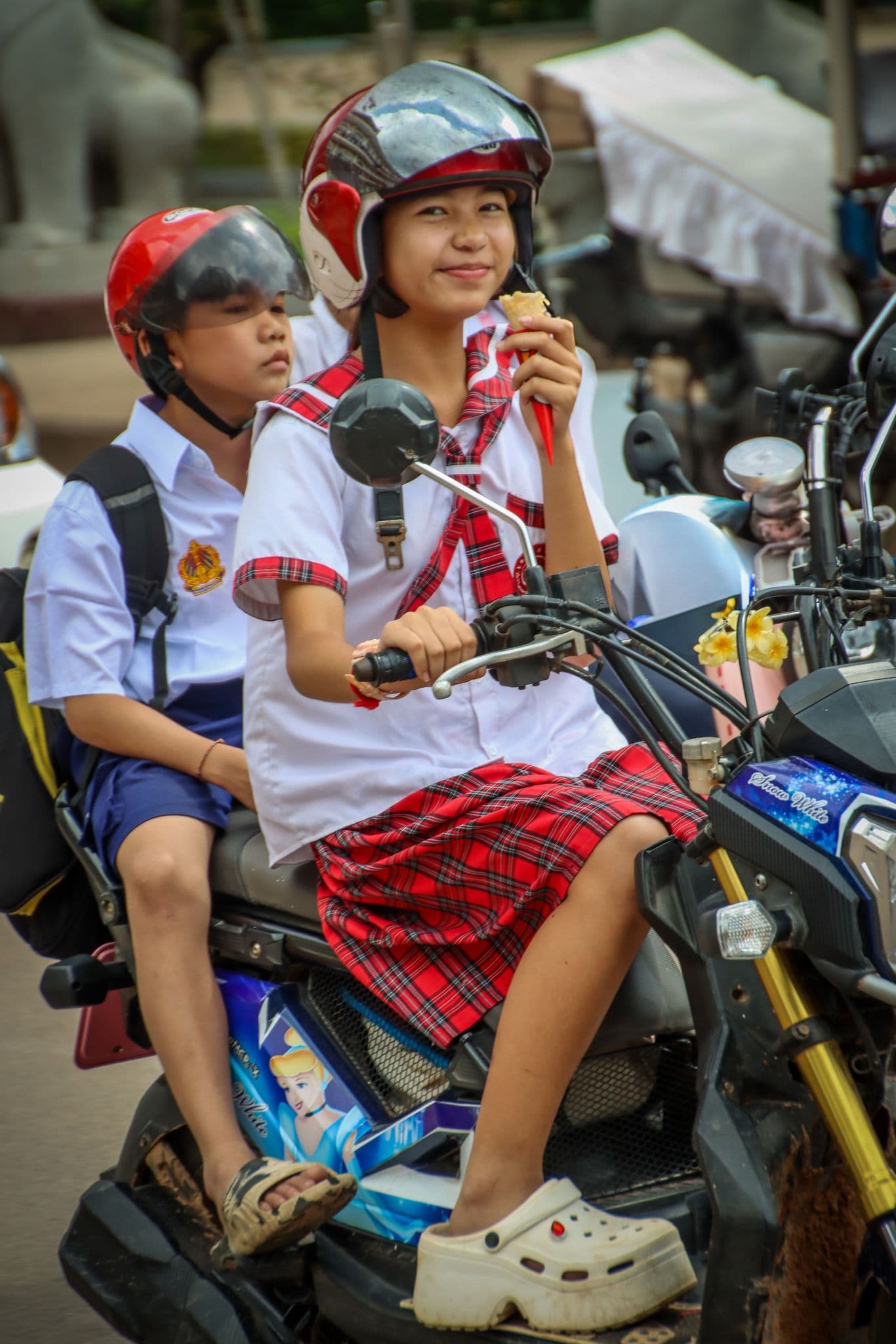 Two students riding a motorbike on a busy street in downtown Siem Reap, Cambodia, with one holding an ice cream cone.