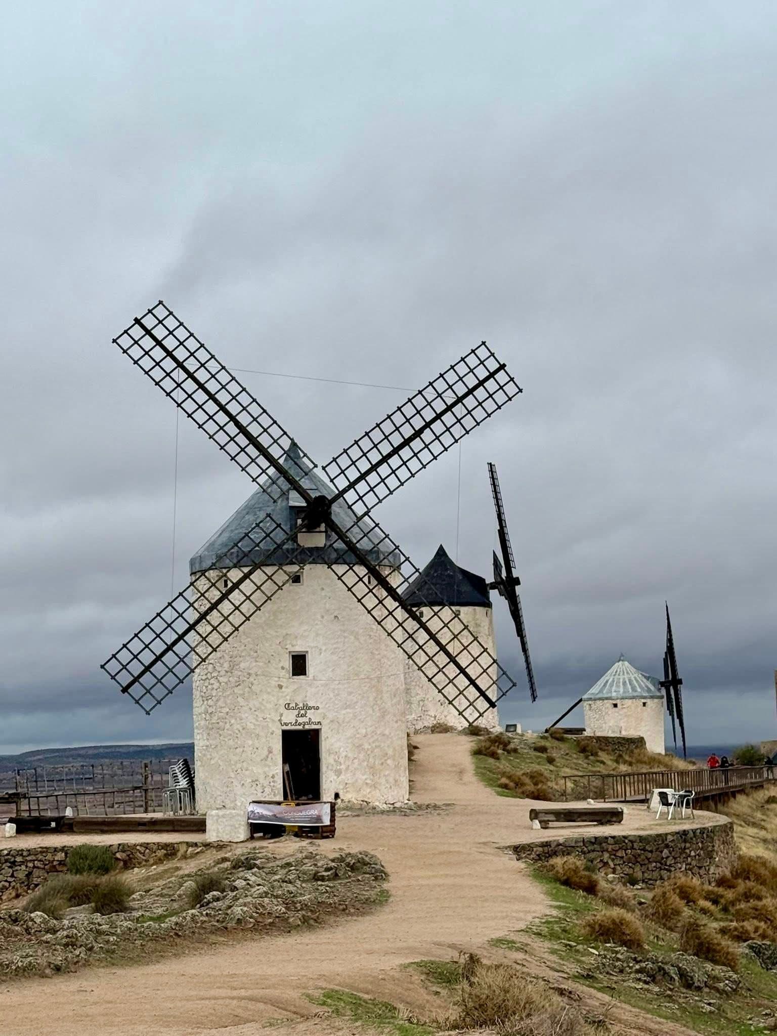 White stone windmills on a grassy hill in La Mancha, Spain with a dirt path and cloudy sky.