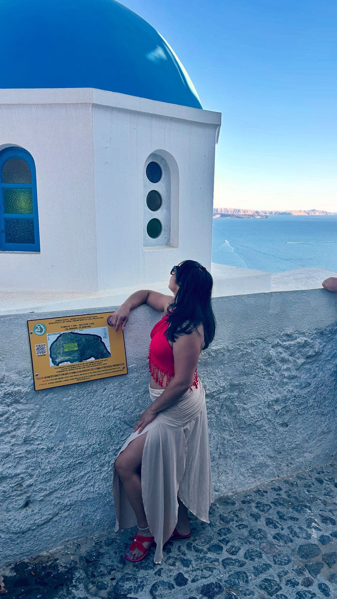 Blue-domed church in Oia, Santorini, Greece with a woman leaning on a white wall and looking toward the Aegean Sea.