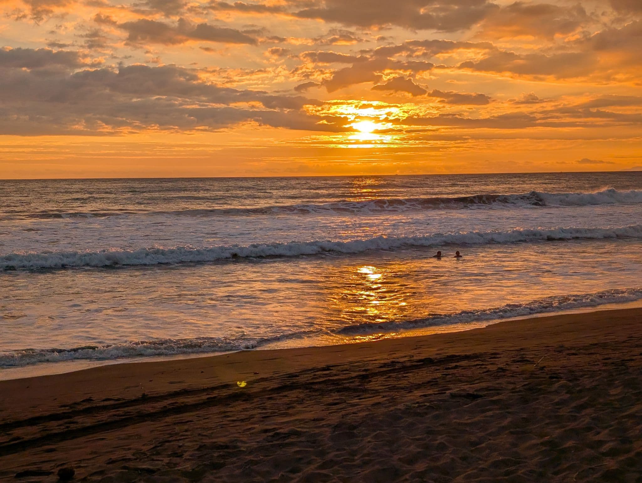 Sunset over Jaco Beach with golden clouds reflecting on the ocean and two people swimming near the shore, Jaco, Costa Rica.