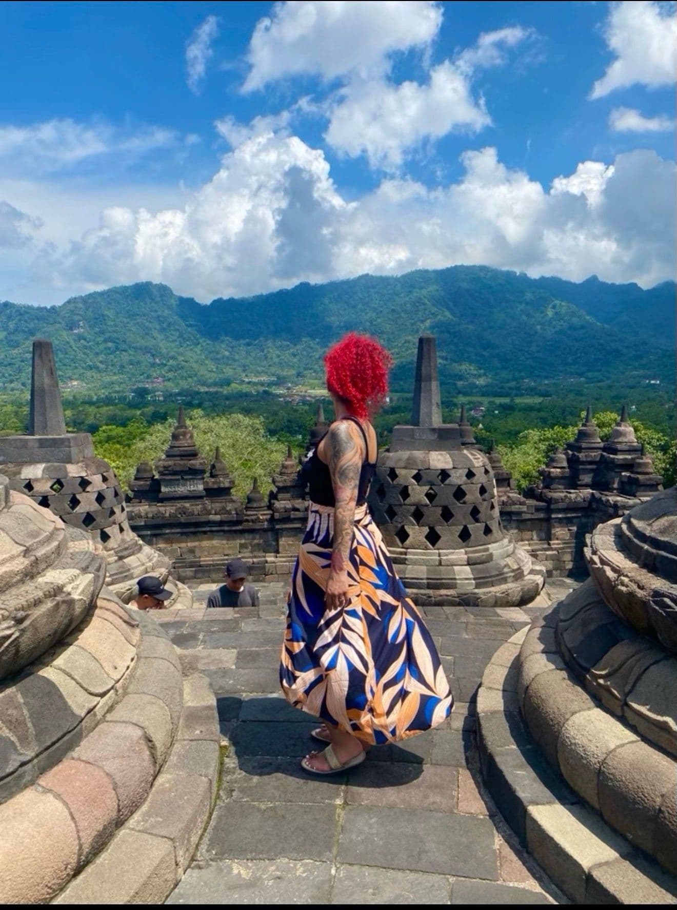 Borobudur Temple stone stupas with a traveler in a patterned skirt standing and looking toward green hills near Yogyakarta, Indonesia.