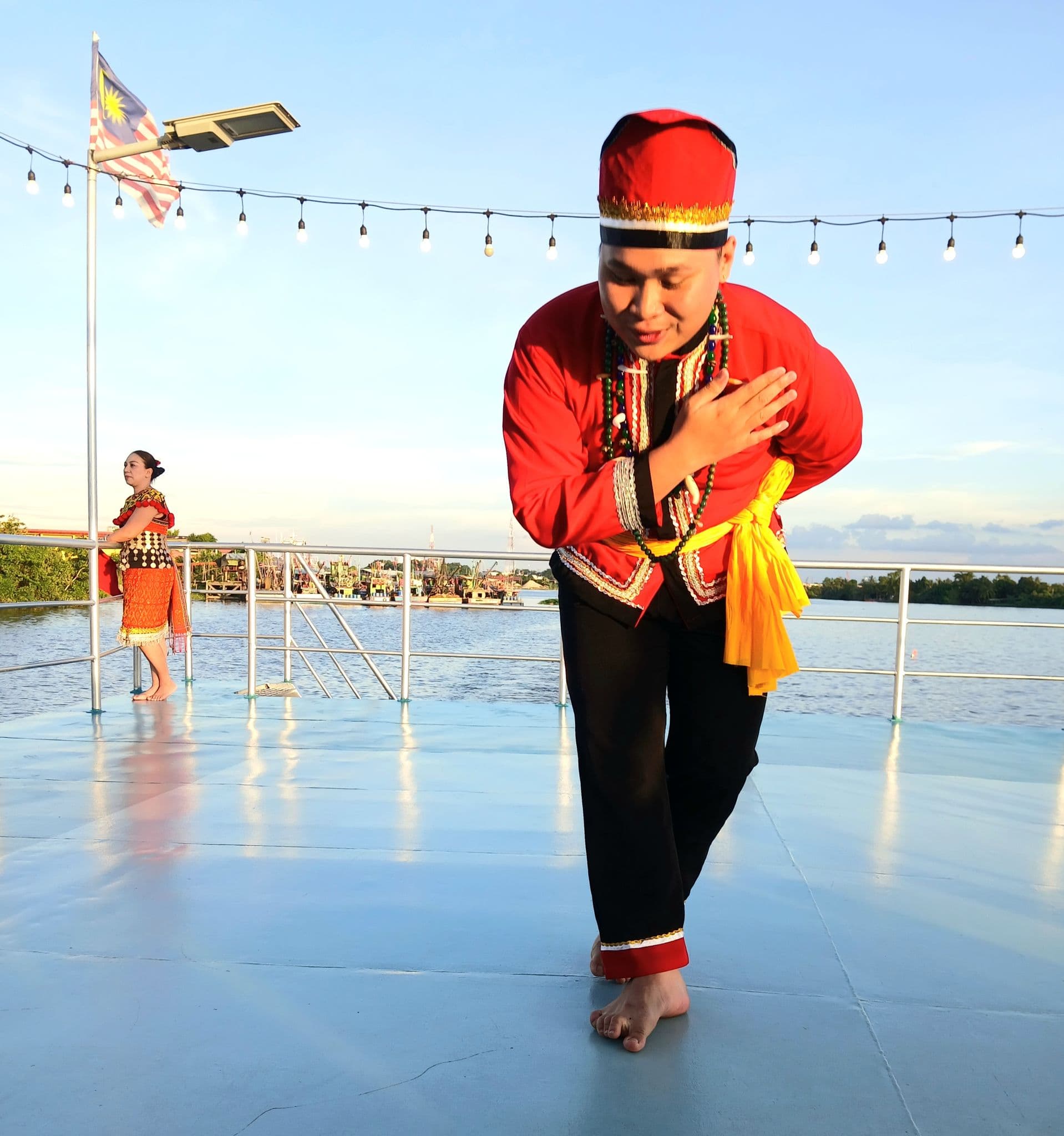 Male dancer in a red traditional costume bowing on a boat deck by the water with a Malaysian flag visible, Borneo, Malaysia.