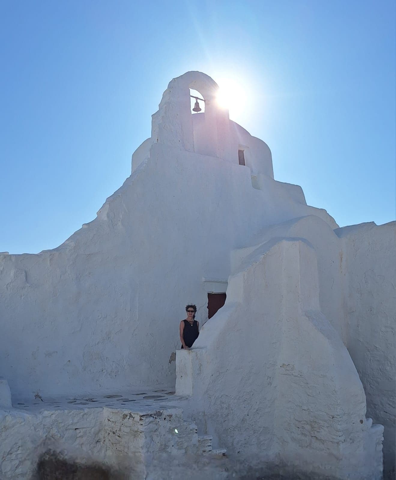Panagia Paraportiani church with the sun behind its bell and a traveler at a whitewashed wall, Mykonos, Greece.