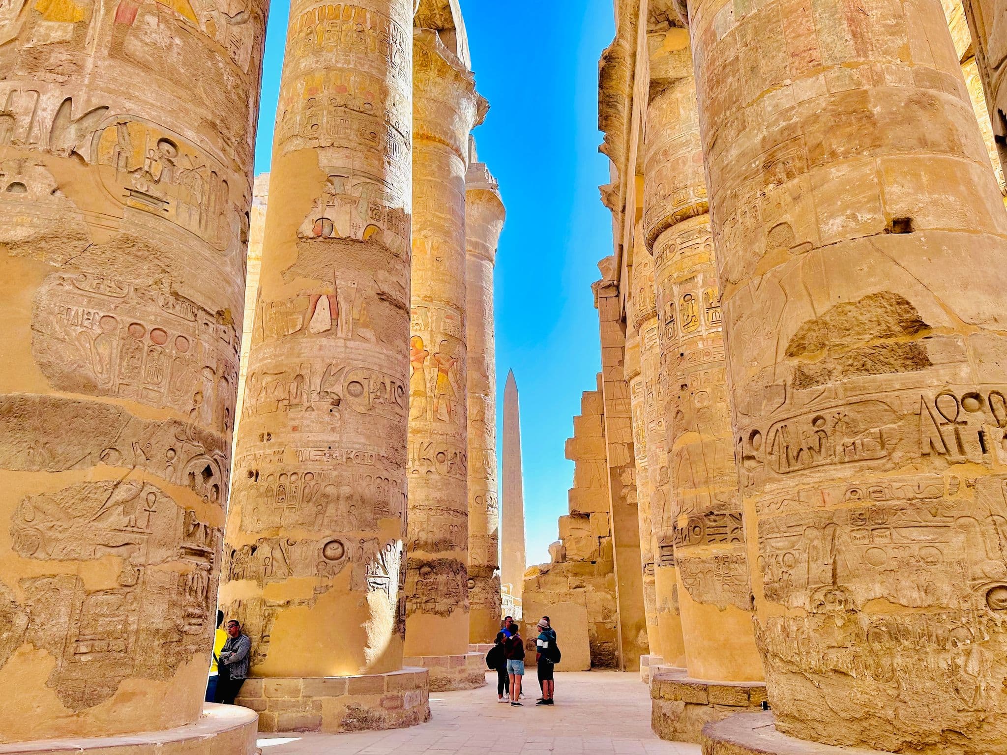 Karnak Temple's massive carved sandstone columns with visitors standing and walking between them and an obelisk visible in Luxor, Egypt.