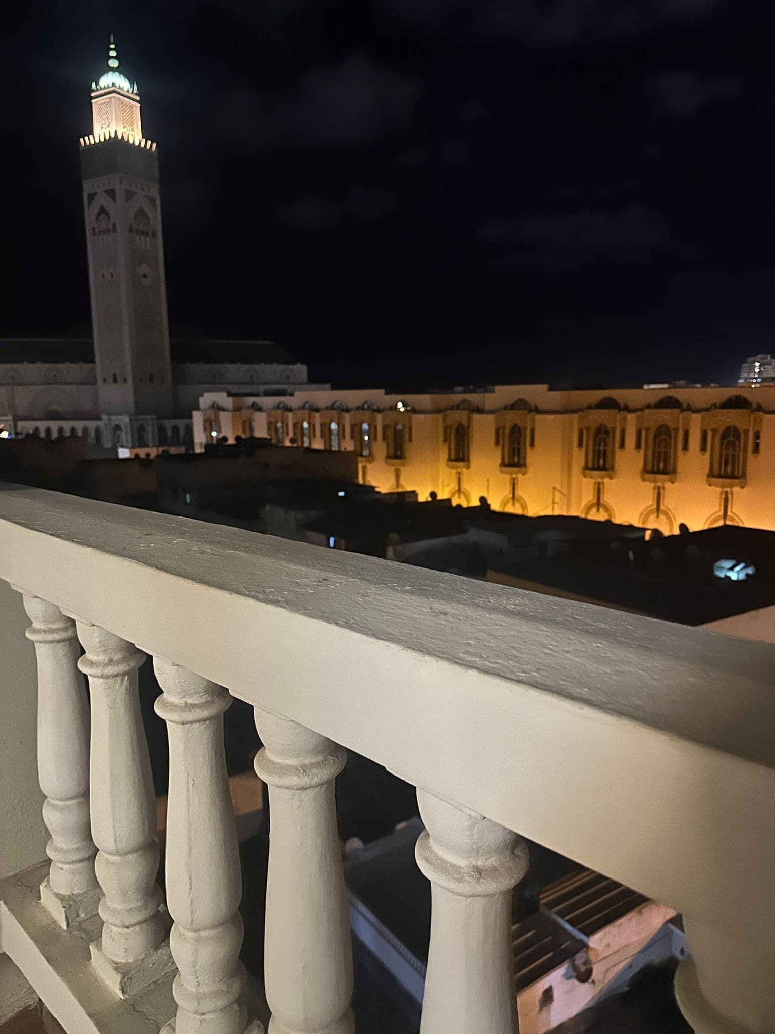 Hassan II Mosque lit at night with a white balcony railing in the foreground on a hotel balcony, Casablanca, Morocco.
