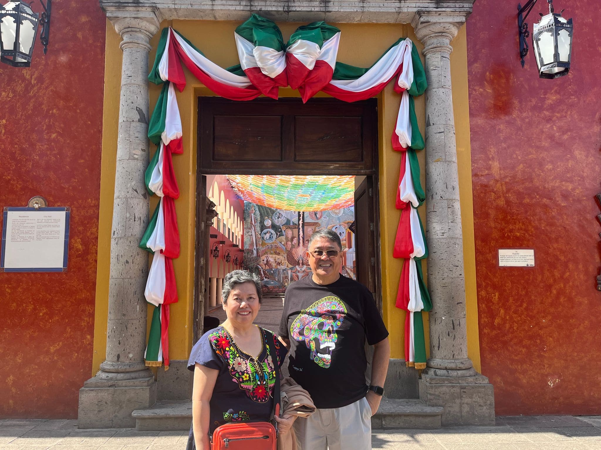 Doorway draped in green, white, and red fabric with two travelers posing in front and a painted courtyard visible inside, Mexico.
