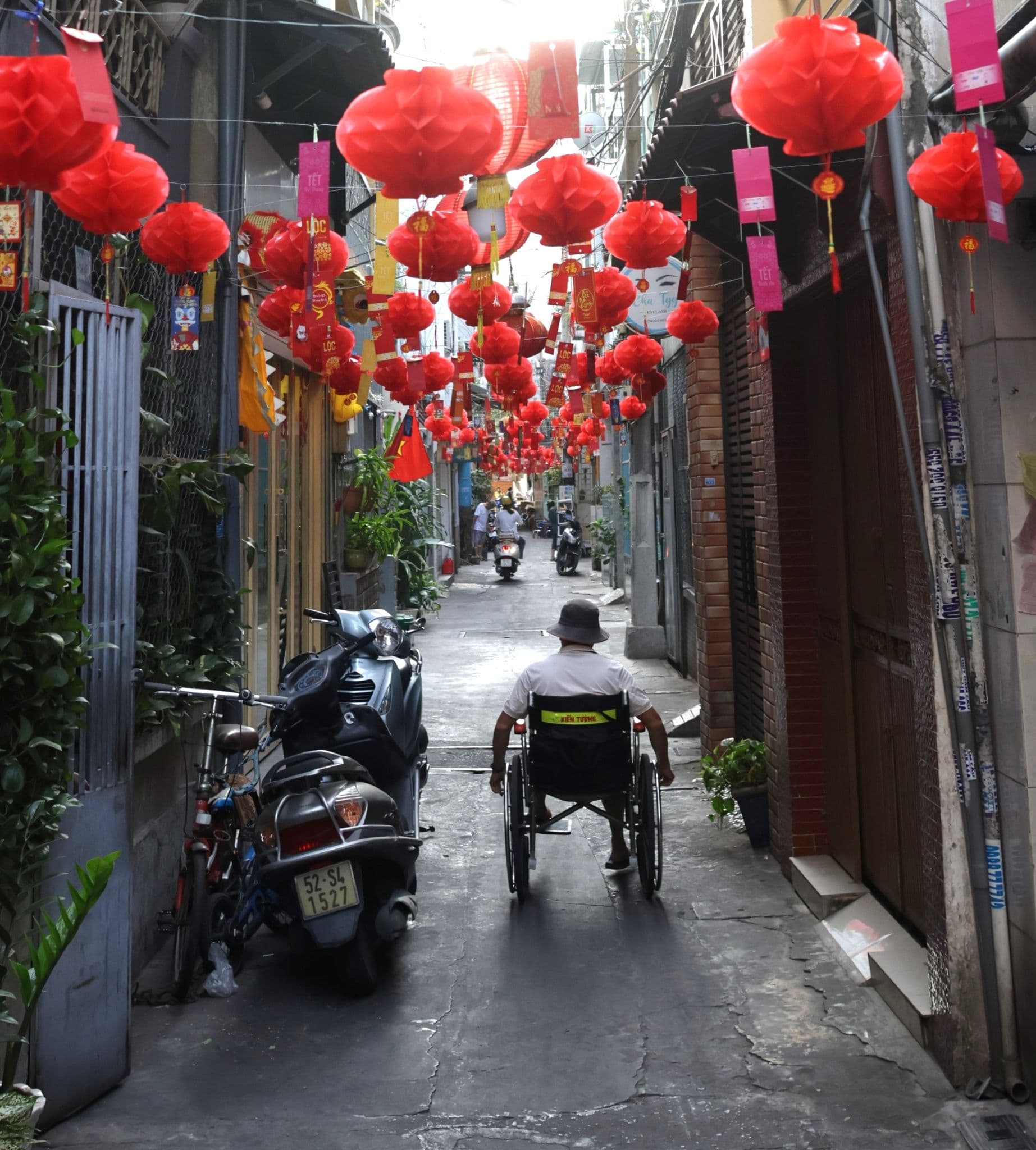 Red lanterns strung over a narrow alley in Ho Chi Minh City, Vietnam, as a person in a wheelchair rolls past parked scooters.