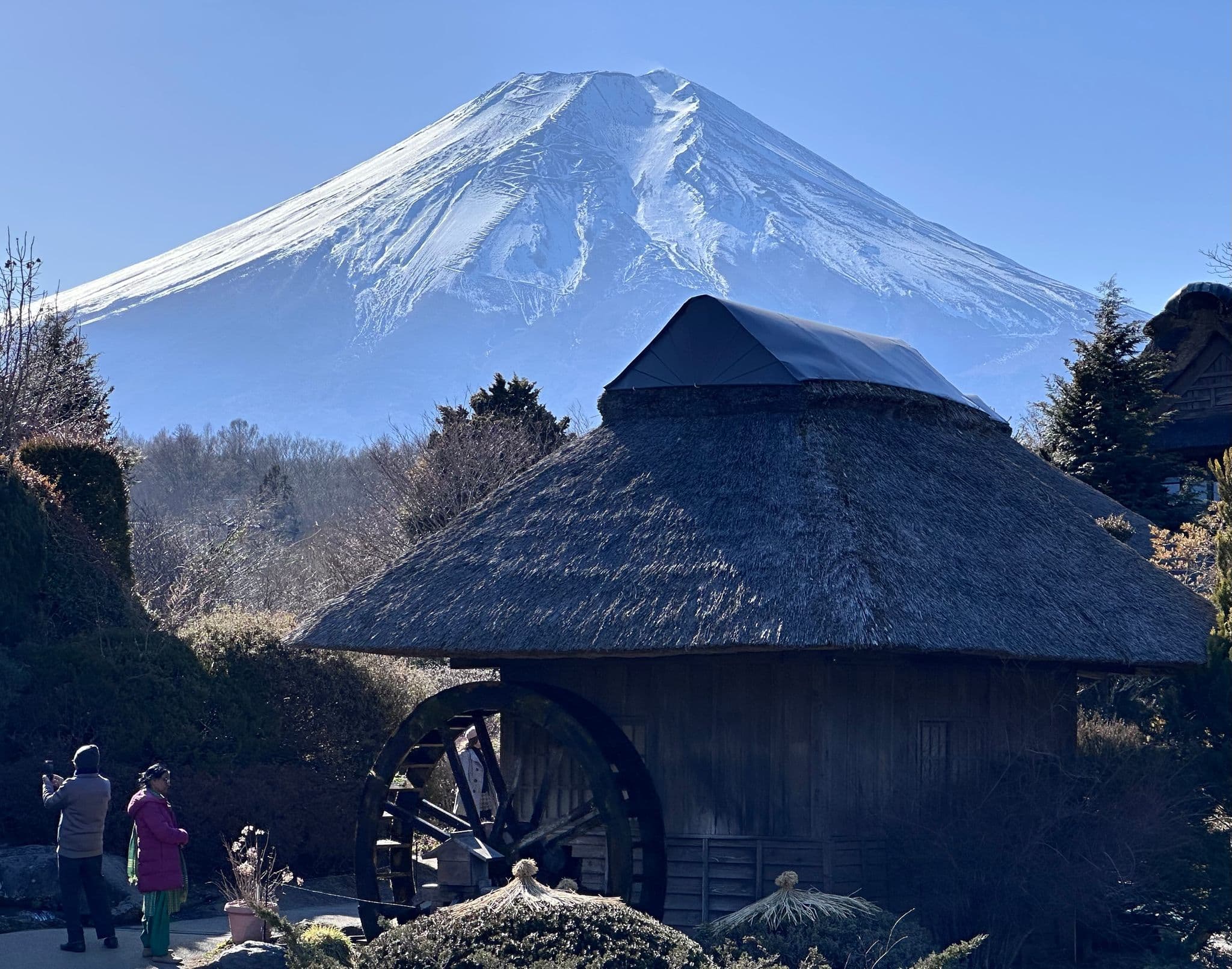 Mount Fuji rising behind a thatched-roof building with a water wheel and visitors in Oshino, Japan.