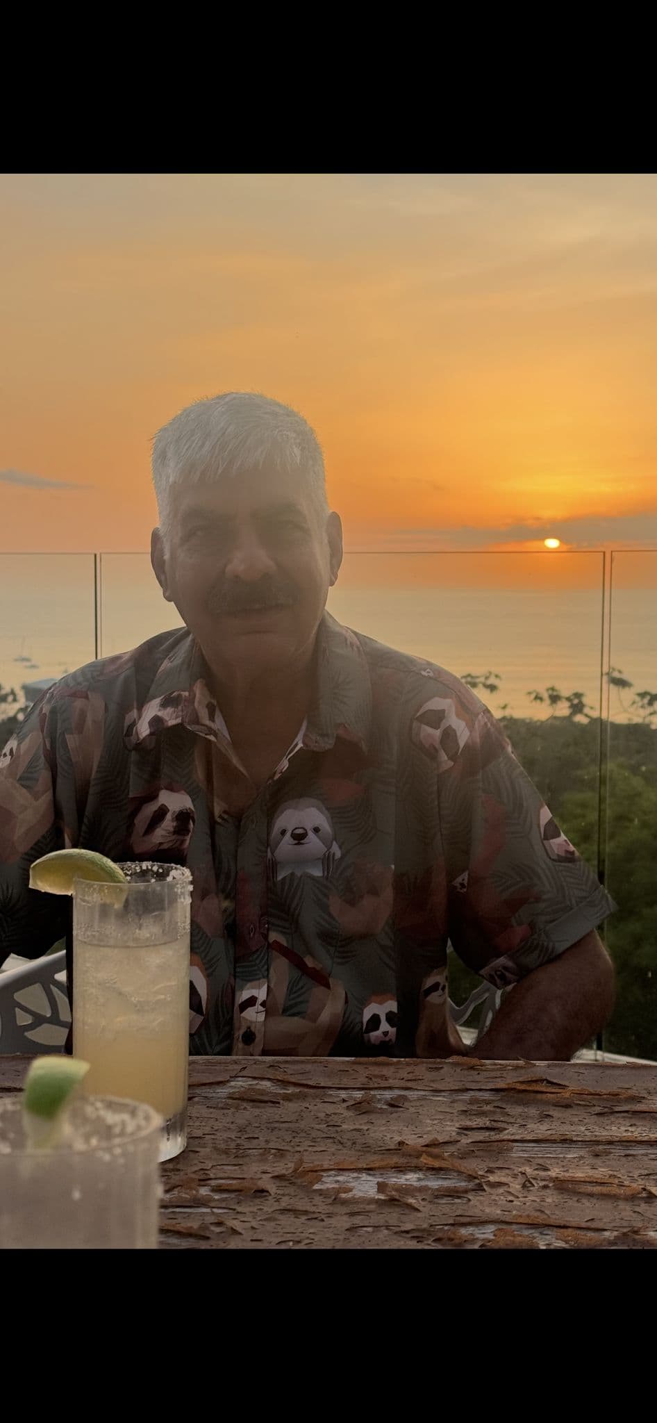Sunset over the ocean with a man in a patterned shirt seated at a balcony table with salt-rimmed drinks.