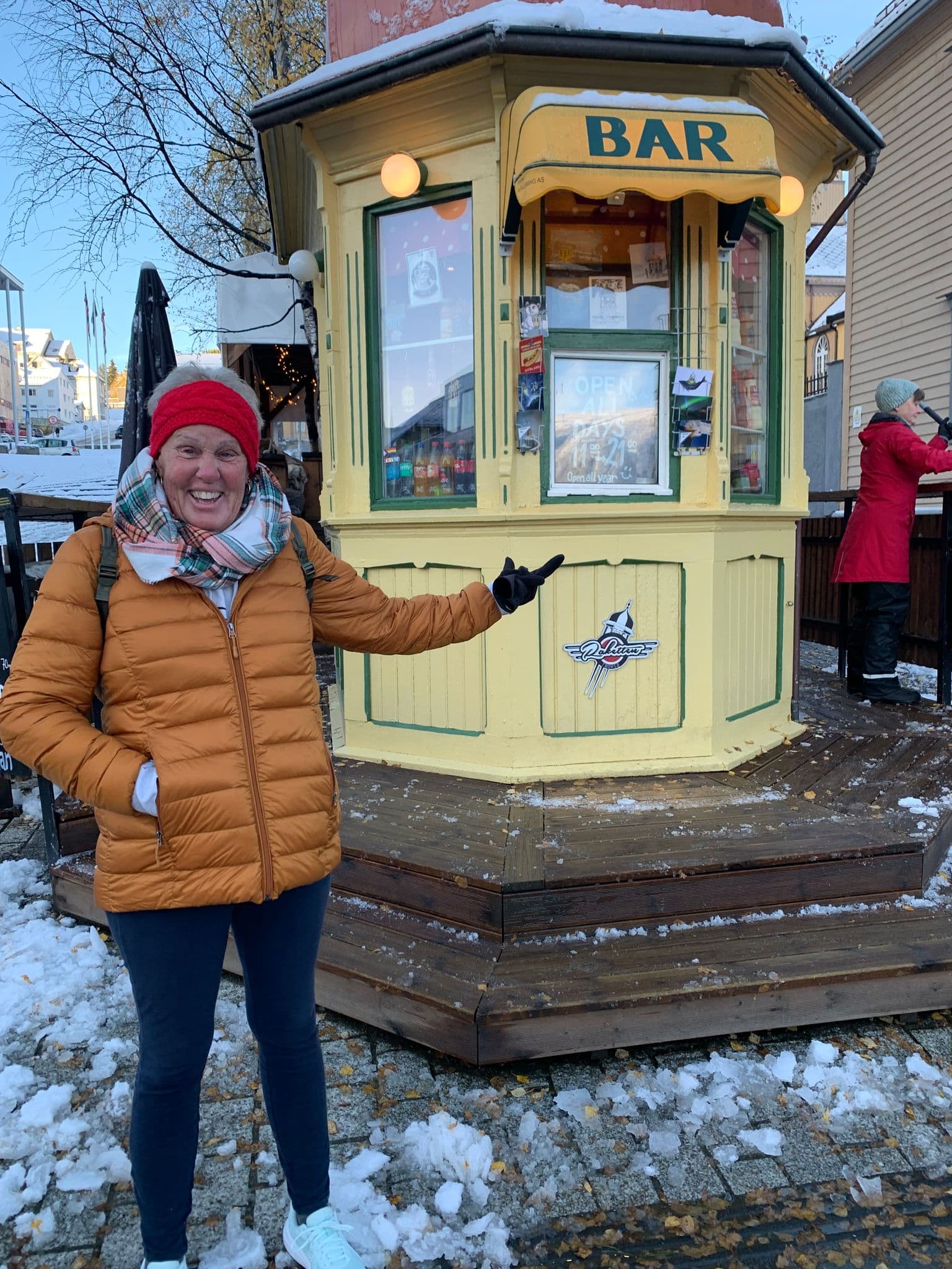 Small yellow kiosk labeled "BAR" with a smiling traveler pointing at it on a snowy street in Norway.