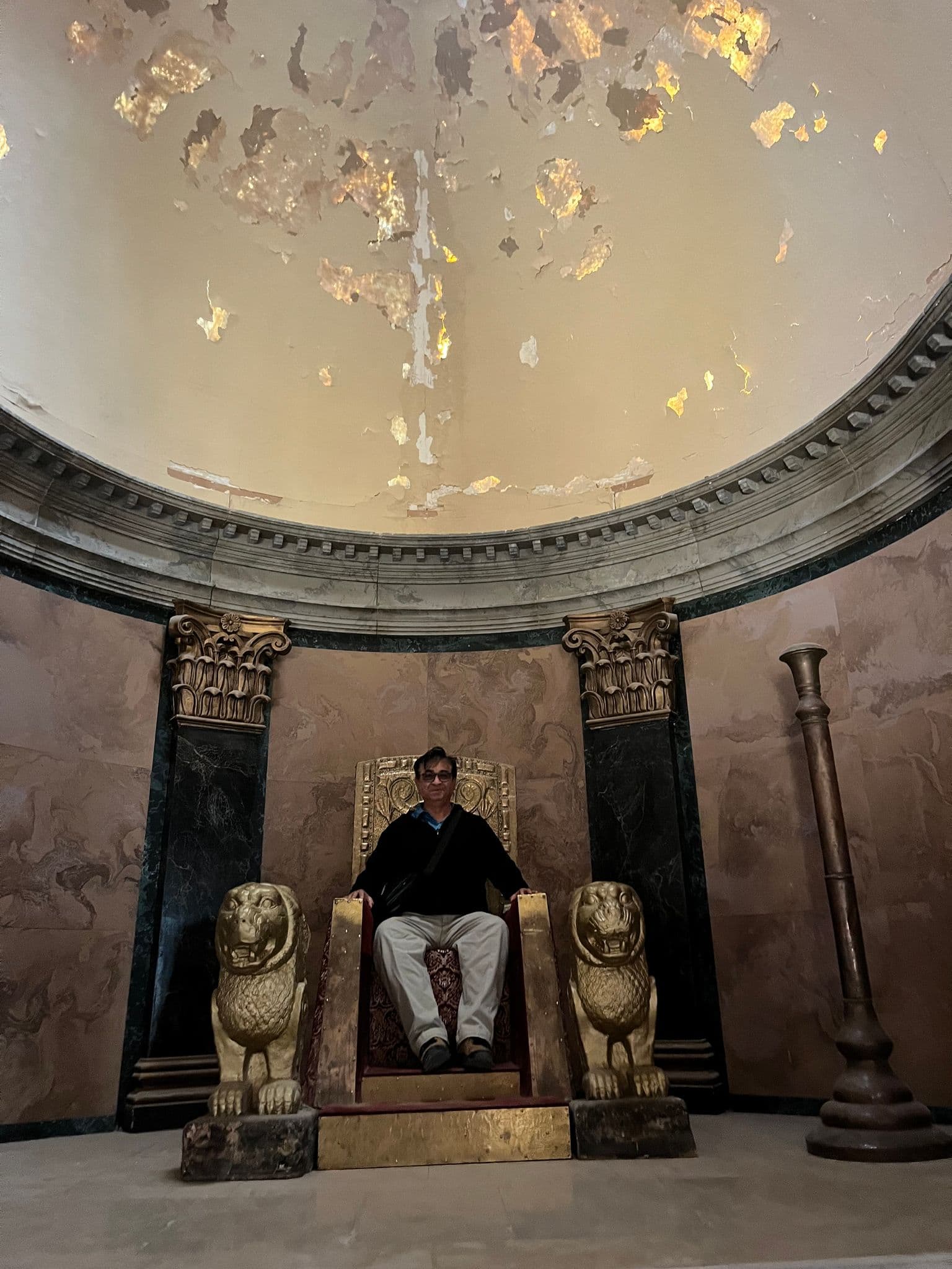 Gilded throne with carved lion armrests and a person seated beneath a peeling domed ceiling in Morocco.