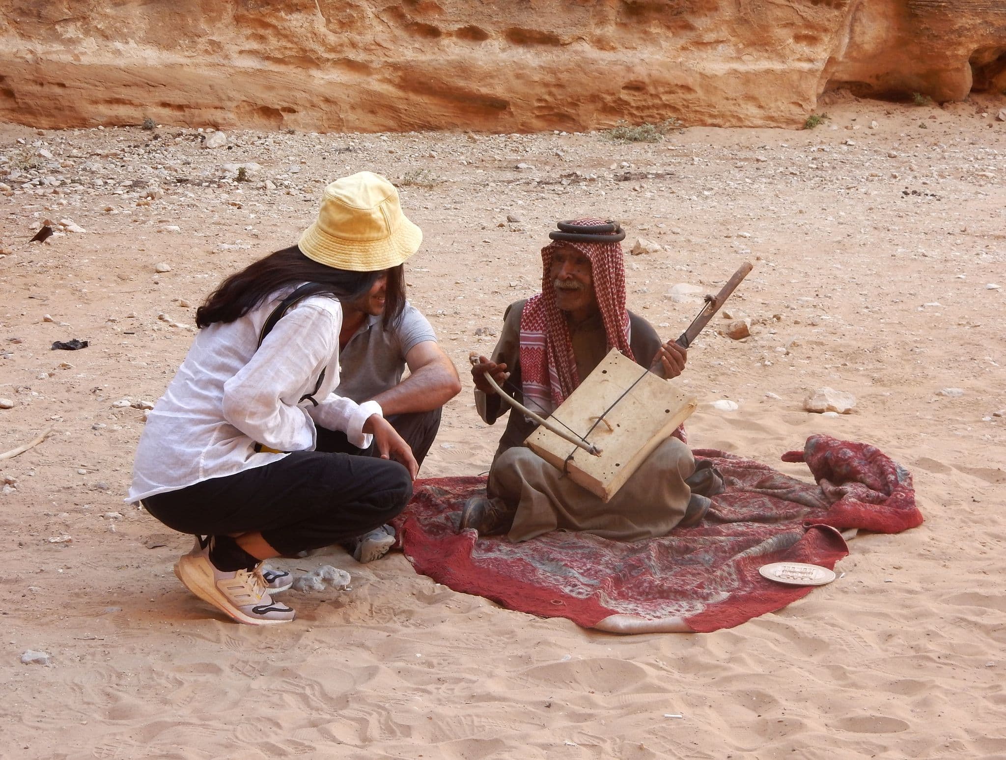 Man playing a traditional bowed instrument on a rug in Little Petra (Siq al-Barid), near Petra, Jordan, while tourists listen.