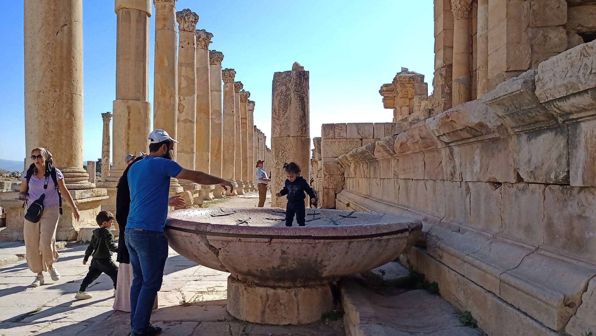 Ancient Roman columns and a large stone basin at Jerash, Jordan, with a child standing in the basin as adults look on.