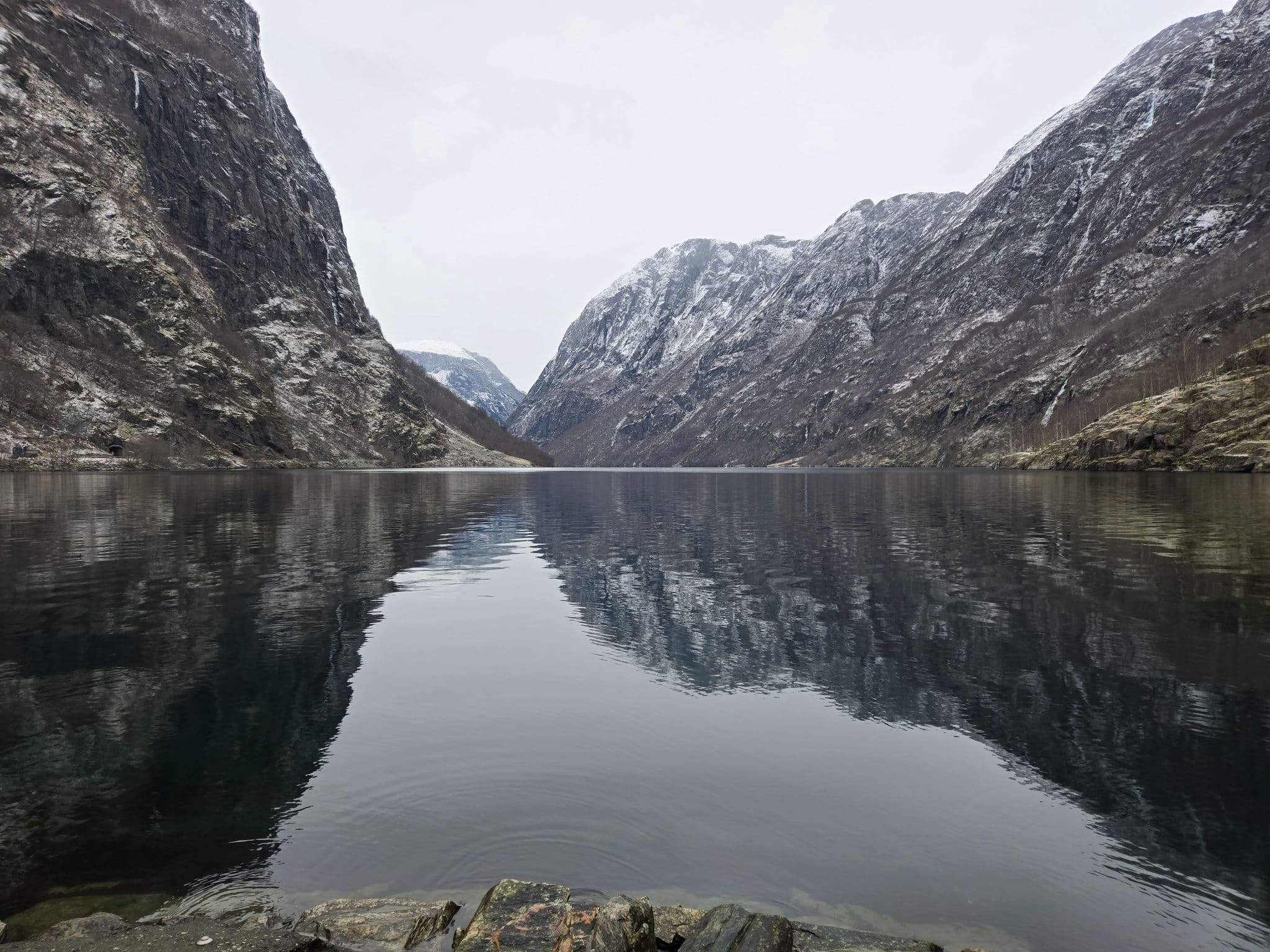 Snow-dusted Norwegian fjord framed by steep rocky cliffs reflected in calm water with shoreline rocks in the foreground, Norway